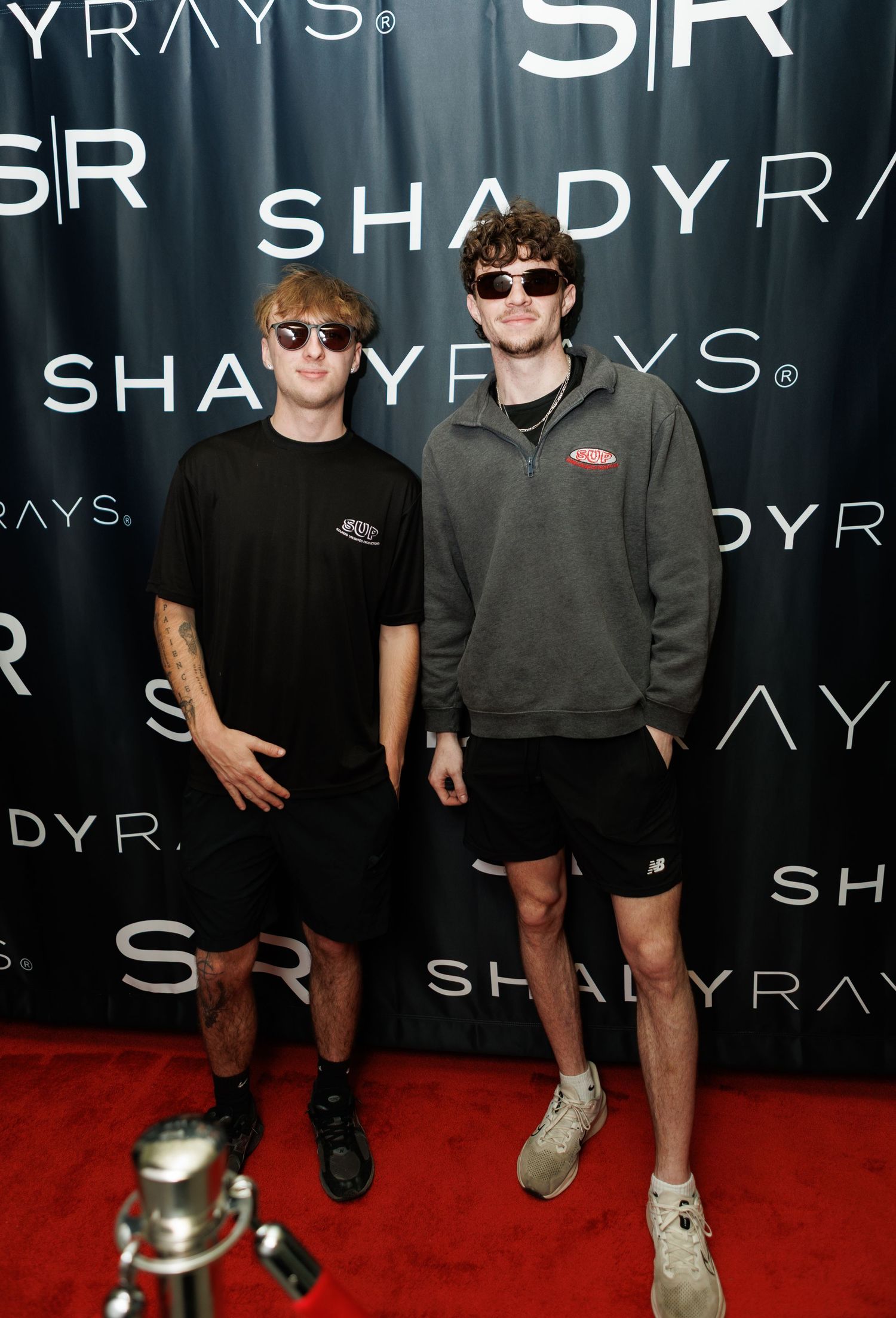 Two young men wearing sunglasses and casual outfits pose on a red carpet at a Shady Rays event.