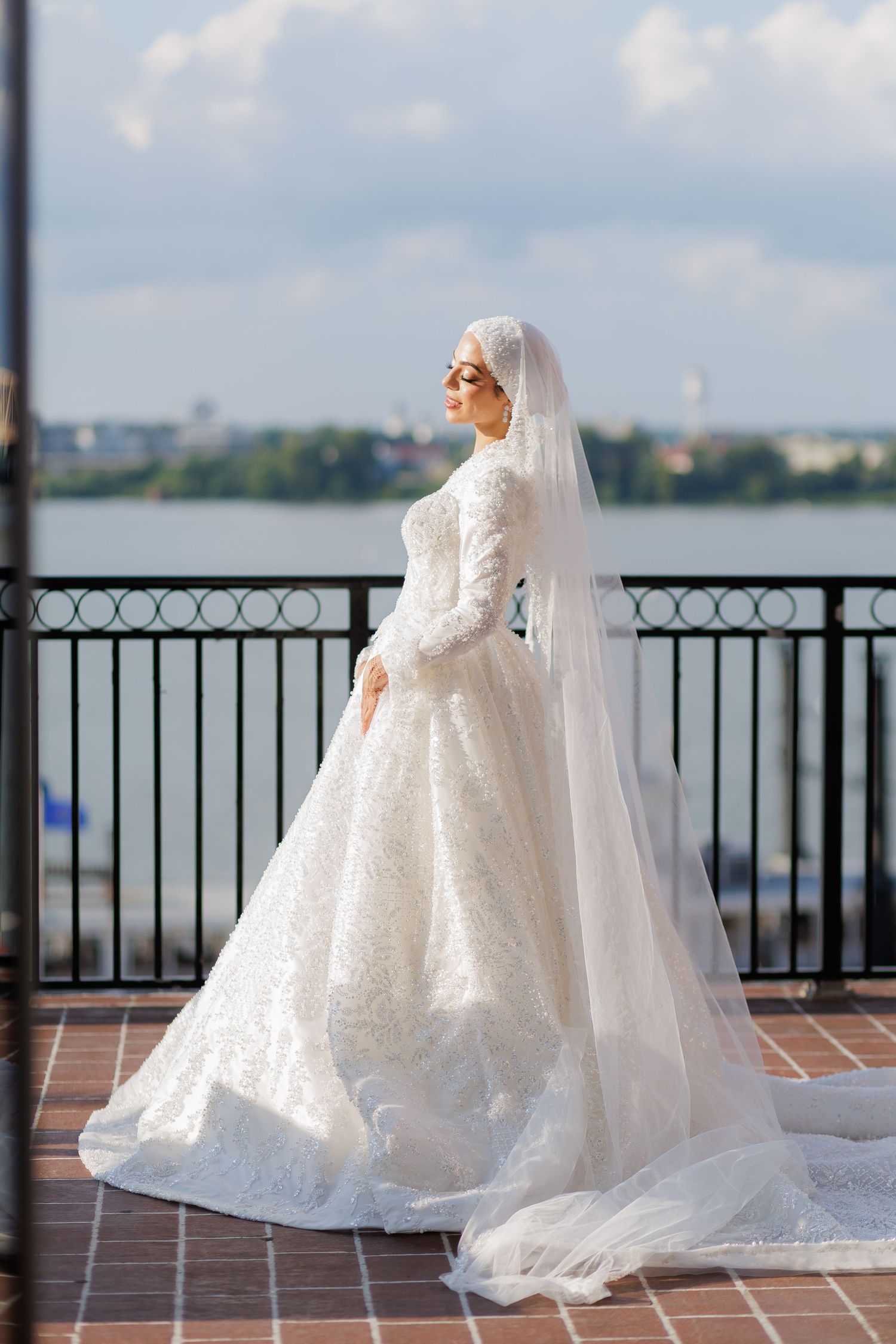 Muslim bride in white lace gown and hijab veil stands on waterfront balcony, posing elegantly on her wedding day.