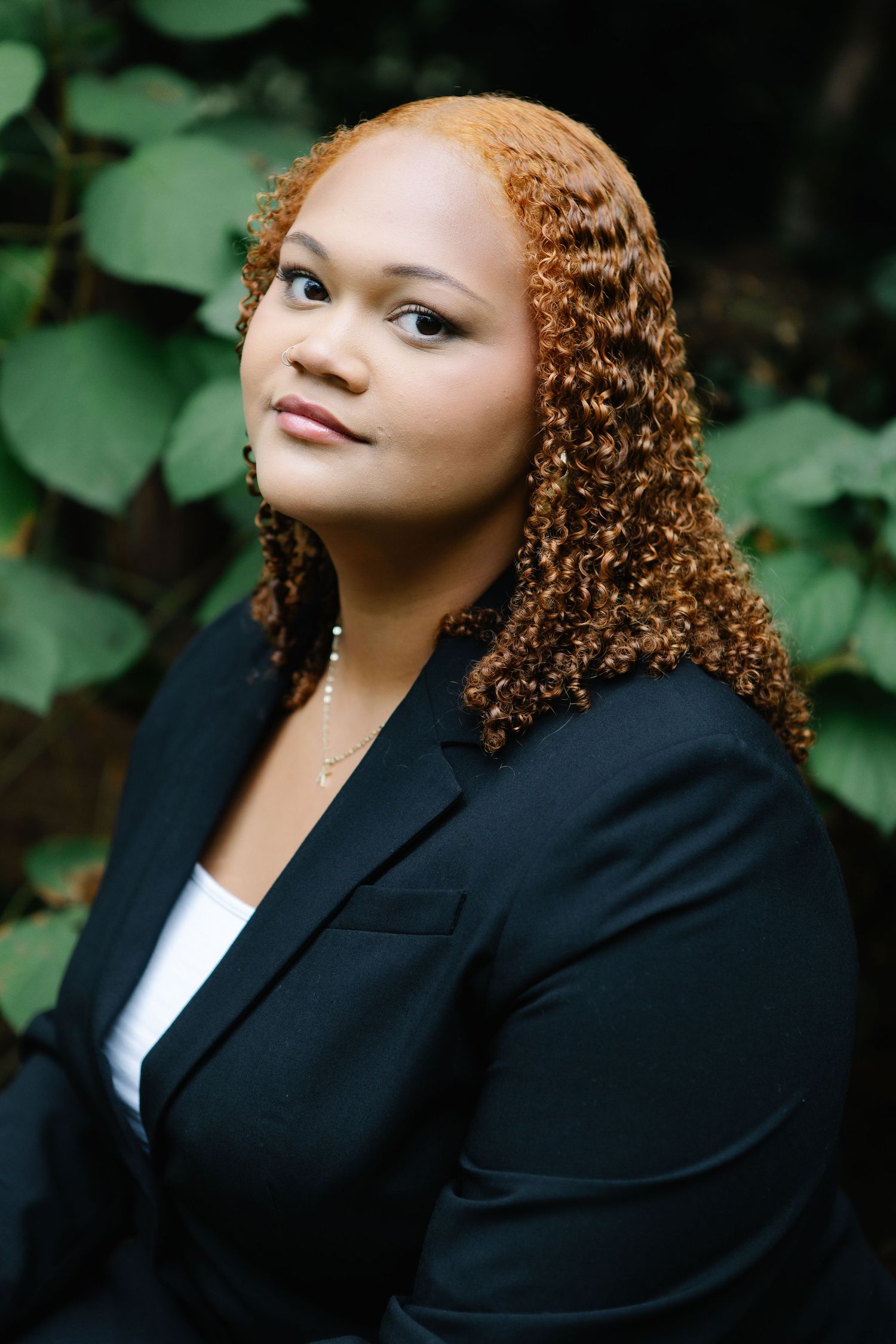 Professional portrait of person in black blazer against green foliage background.
