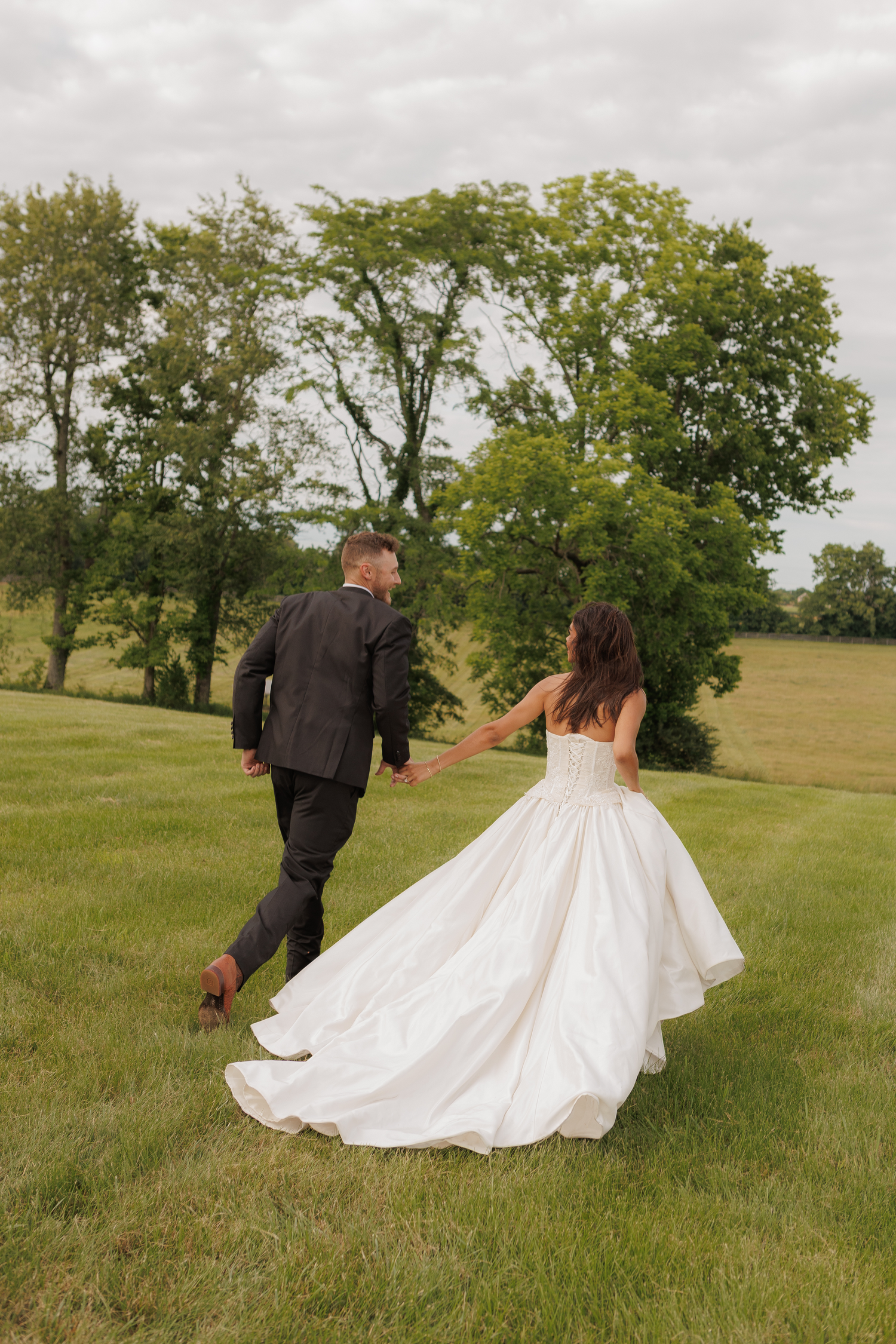 Bride and groom holding hands walking through green field in wedding attire, white ball gown dress and dark suit.
