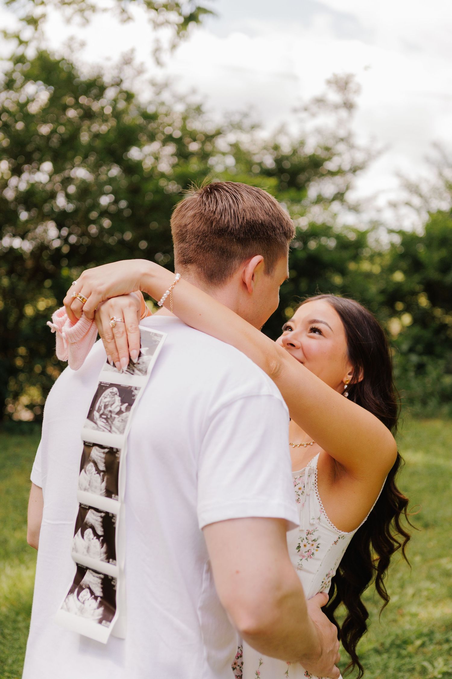 Happy couple embracing outdoors, woman holding ultrasound photos on mans back, pregnancy announcement in green garden.