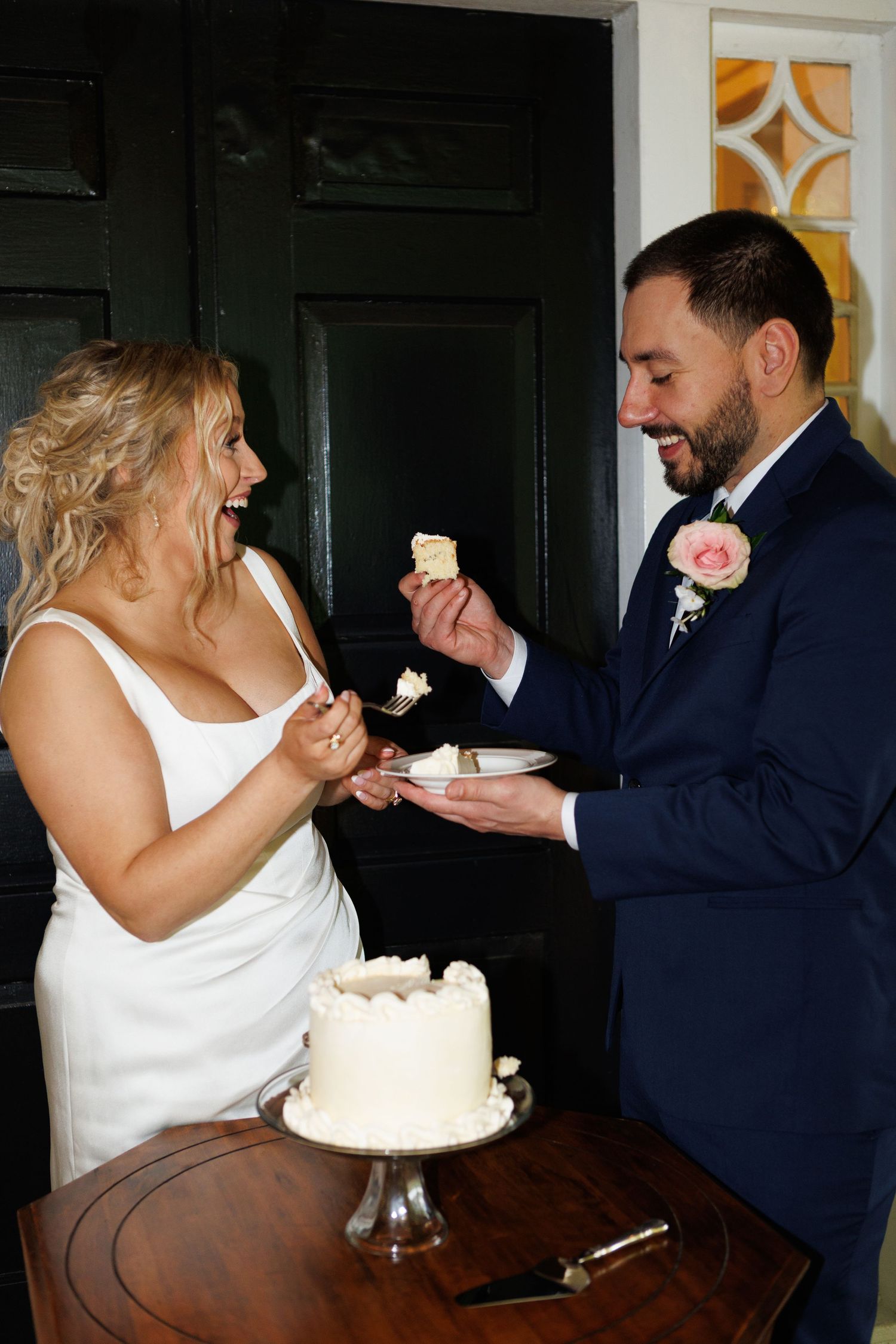 Bride and groom laughing while cutting white wedding cake, groom in navy suit with pink boutonniere.