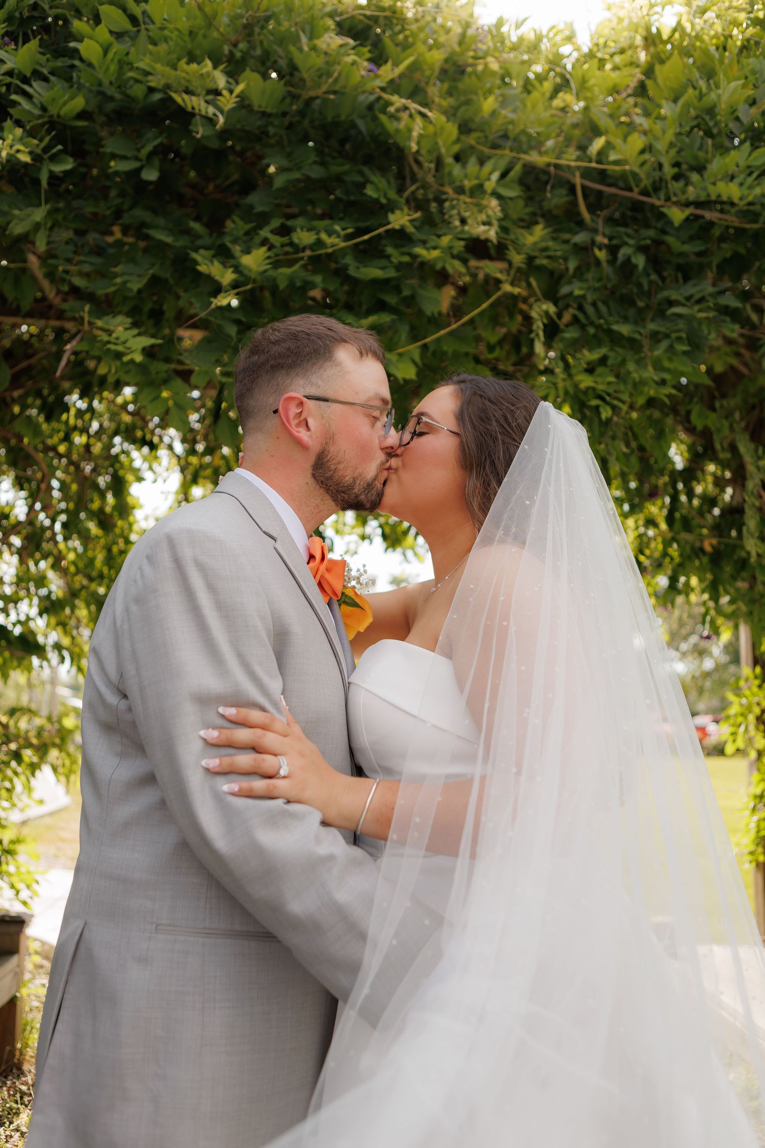 Bride and groom share a romantic kiss under lush green foliage, bride in white gown with veil, groom in light gray suit.