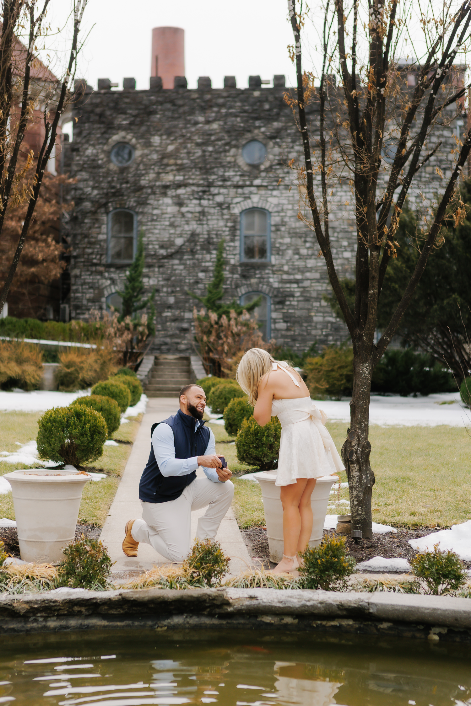 Man proposes on one knee to woman in white dress beside a pond at a stone castle garden.