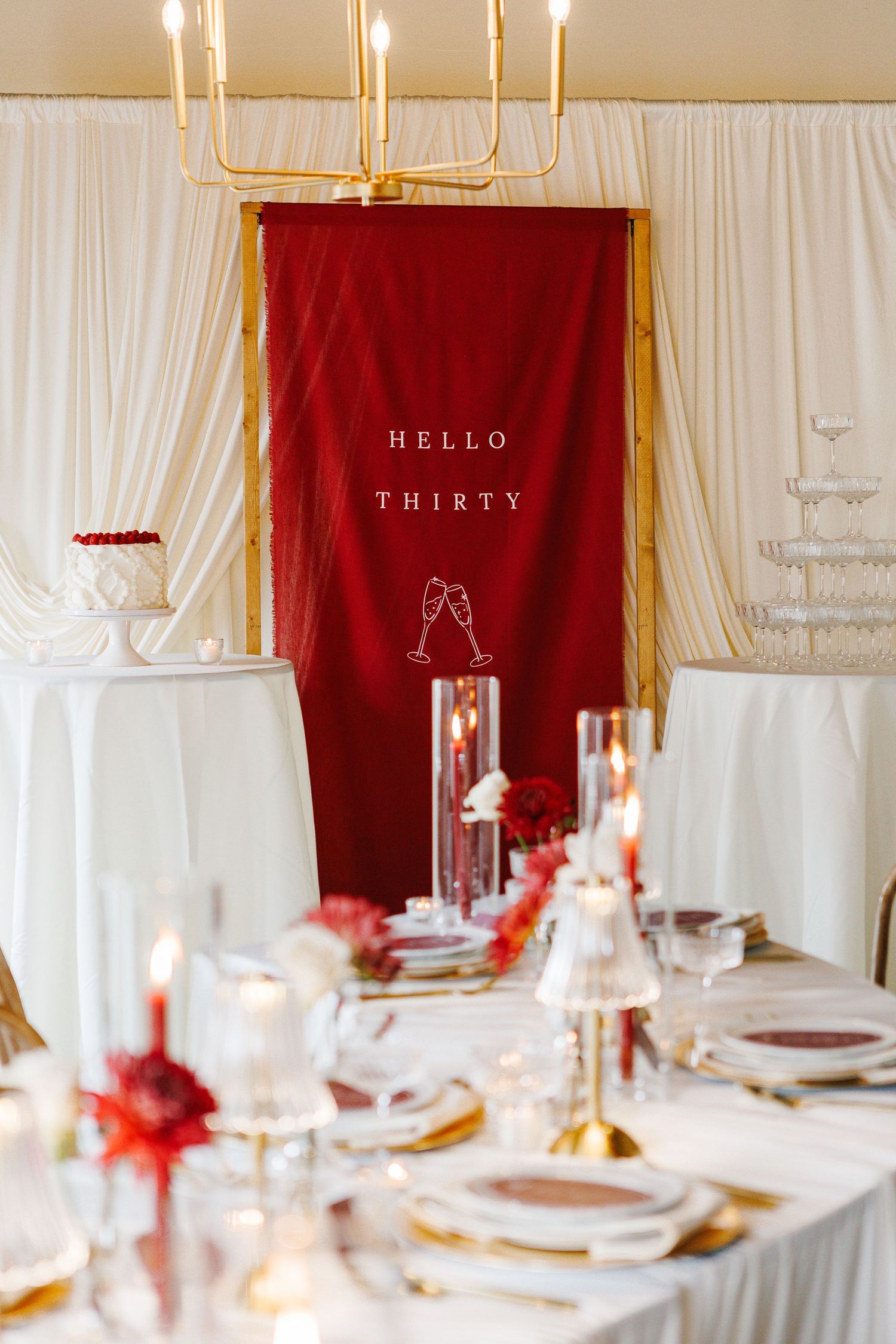 Elegant table setting with white linens and gold candles against red and white backdrop.