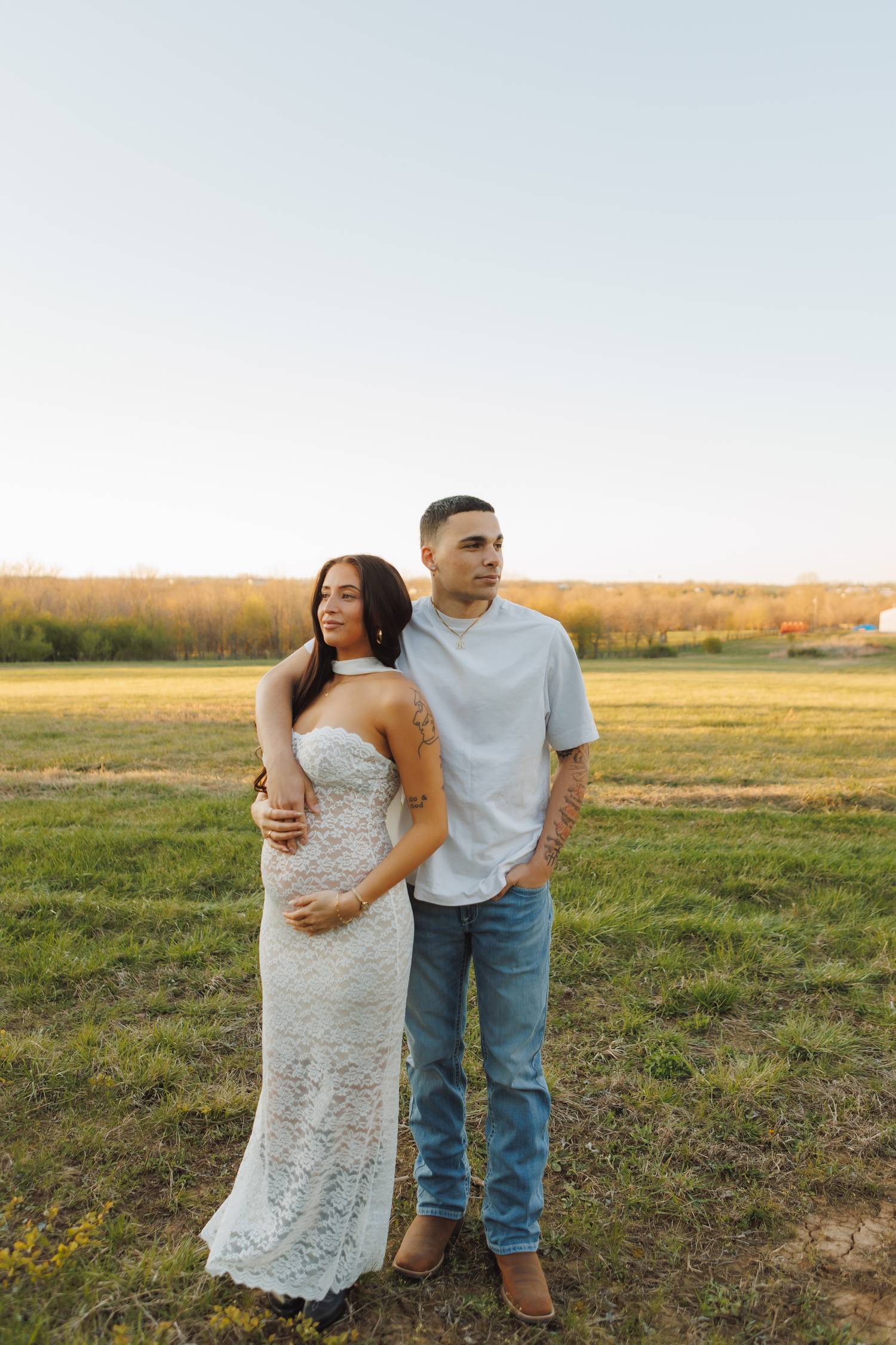 A couple poses in a green field at golden hour, woman in white lace gown, man in jeans and gray tee.