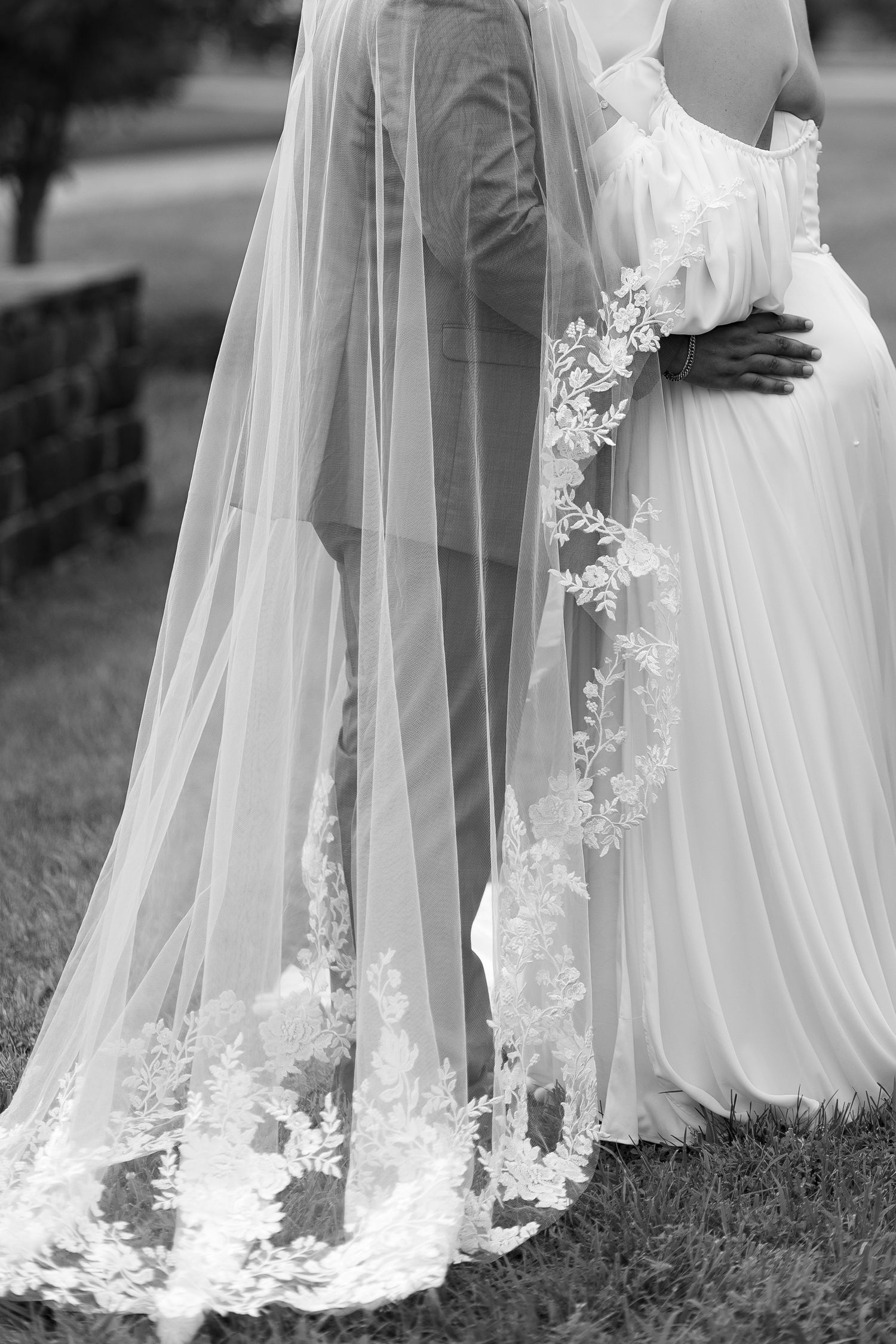 Black and white photo of bride and groom embracing, showcasing elegant lace-trimmed wedding veil trailing on grass.