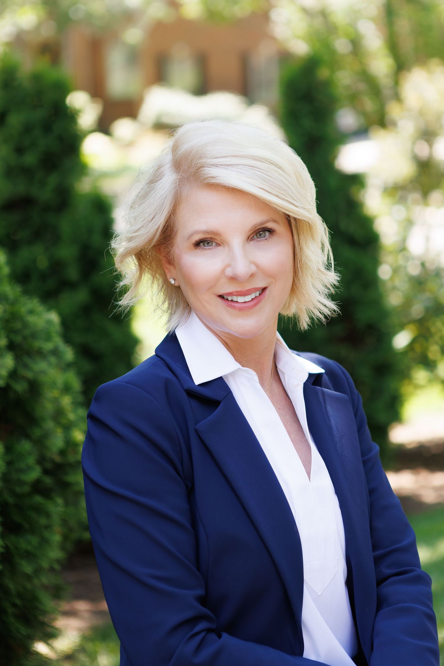 Professional blonde woman in navy blazer smiling outdoors with lush green trees in background.