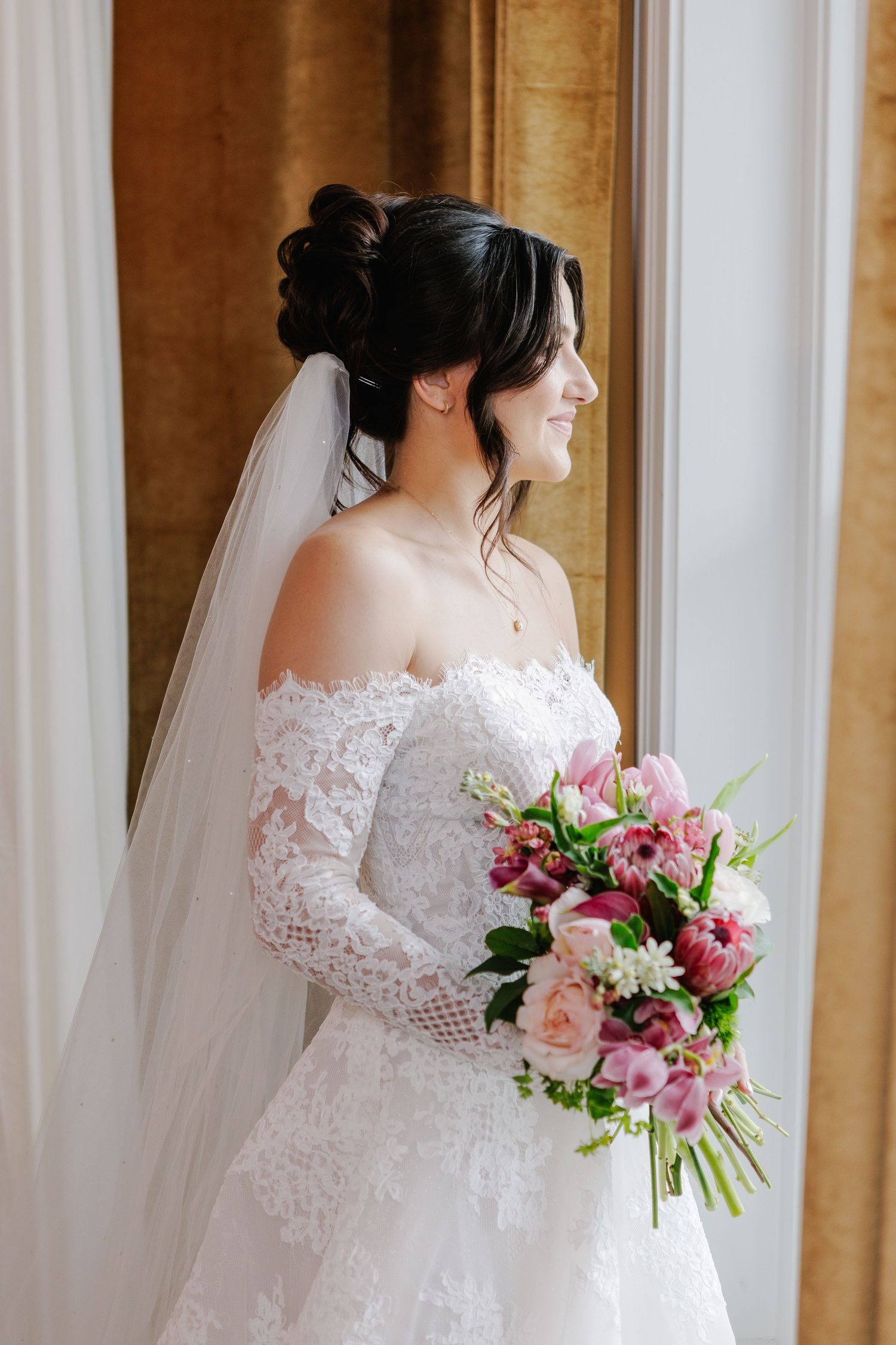 Elegant bride in lace off-shoulder gown holding pink bouquet, standing by window with veil and updo hairstyle.