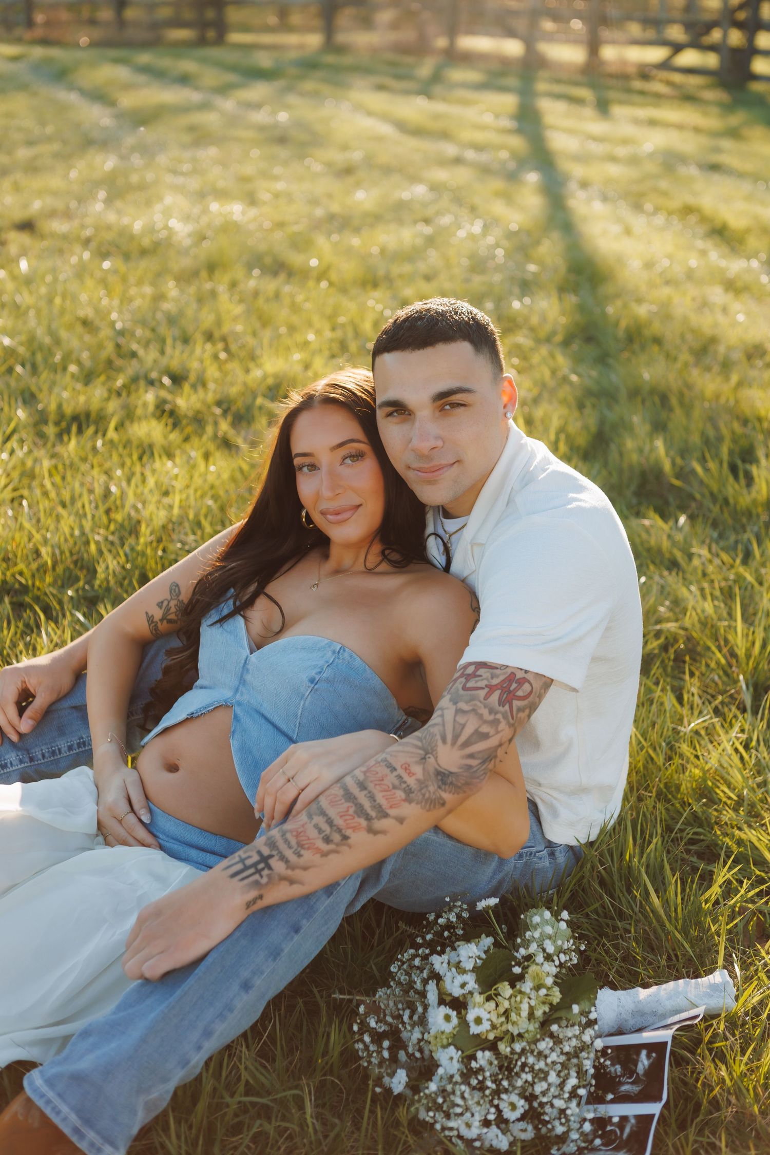 Couple sitting on grass in golden sunlight, pregnant woman in denim top with tattooed partner holding her bump.