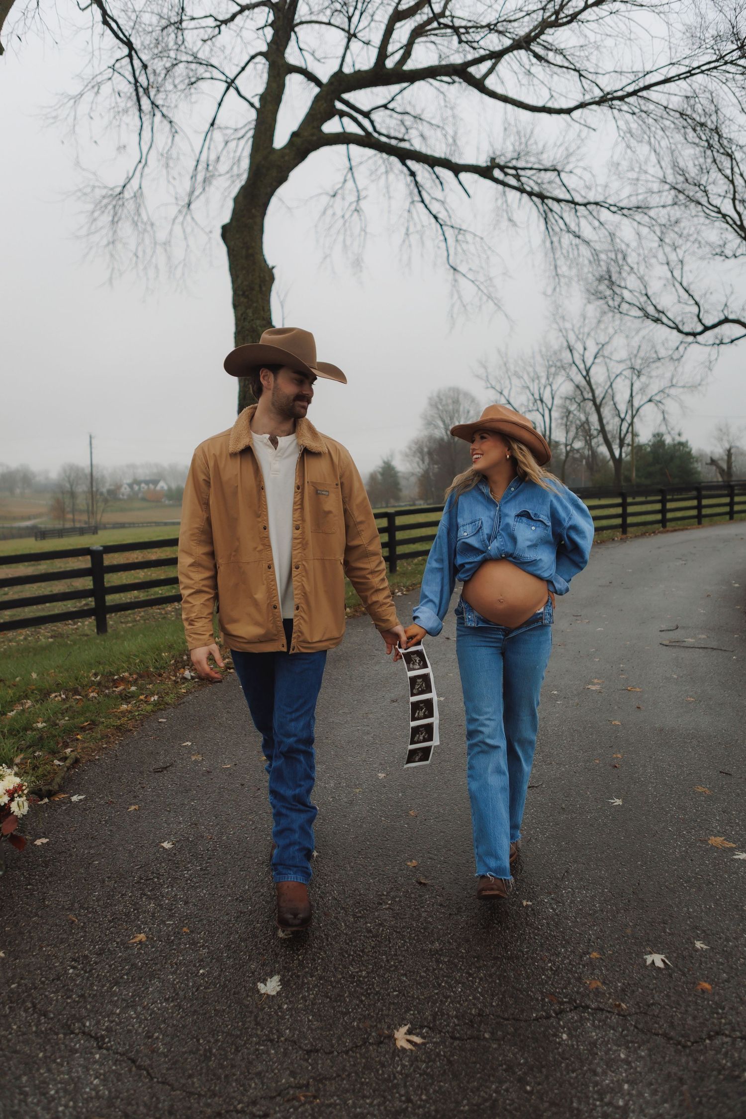 Couple walks down rural path with bare trees and wooden fencing.