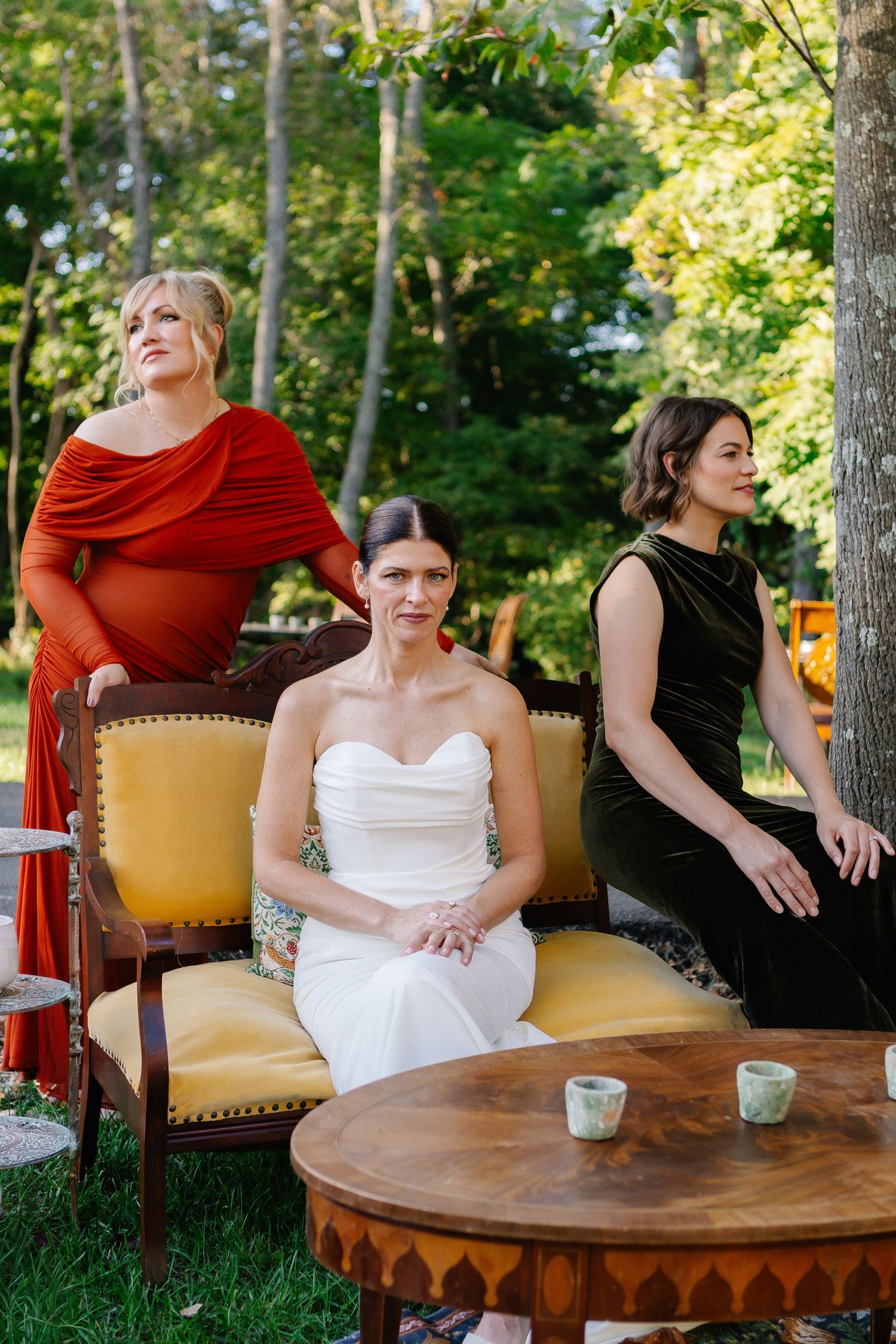 Wedding party relaxing at outdoor table with red accents and natural woodland setting.