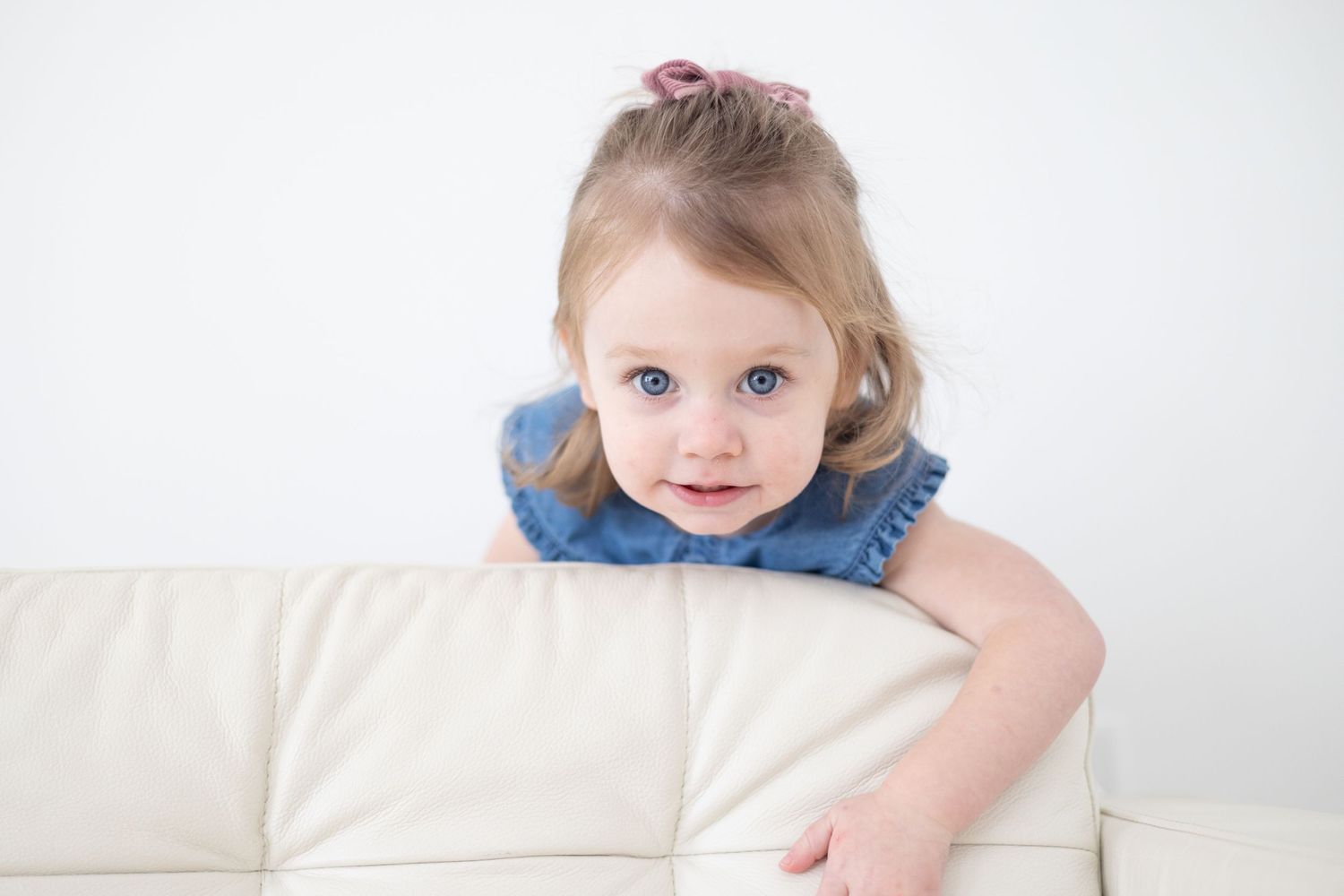 Baby in blue dress resting on white couch with natural lighting.