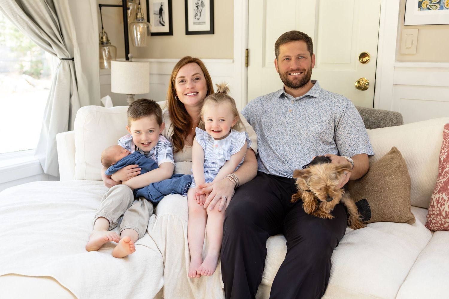 Family of four with two young children and a small dog sitting together on a white sofa in a bright living room.