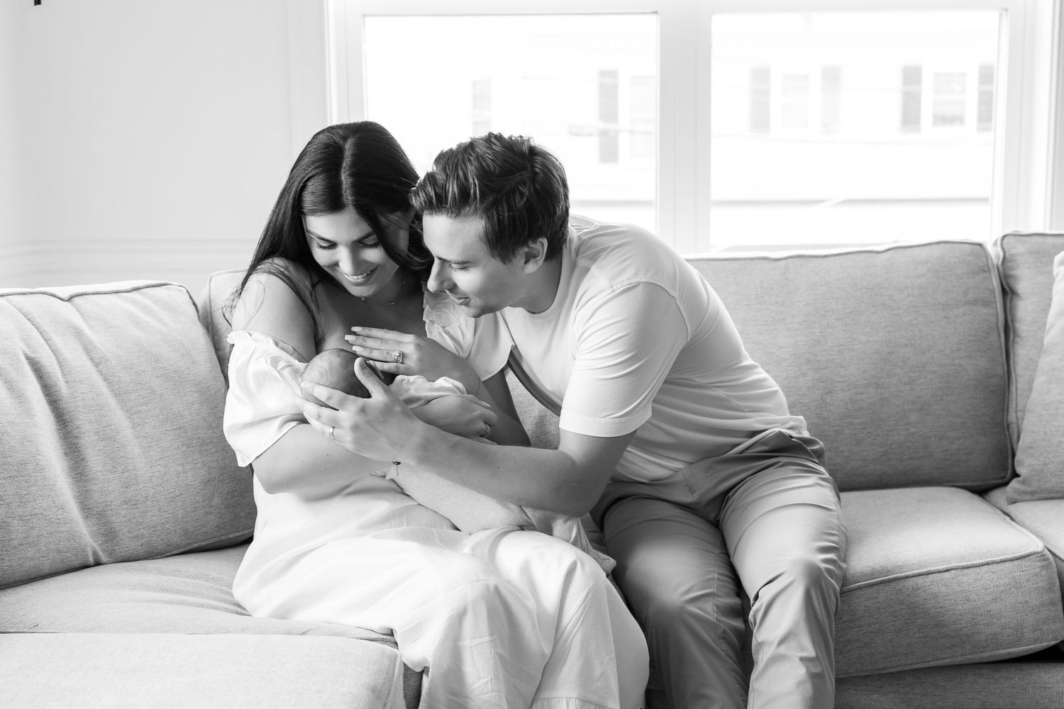 Black and white photo of a couple sitting on a couch, smiling and looking at their newborn baby.