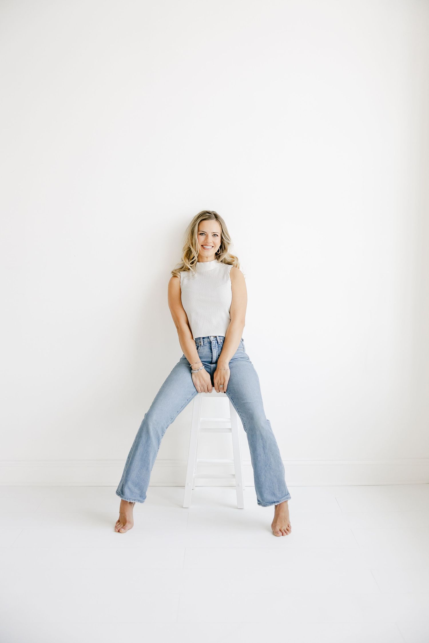 Woman in light blue jeans sitting on white floor in minimalist studio setting.