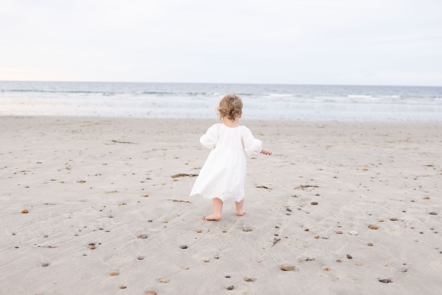Small child in white dress walking along empty beach shoreline.