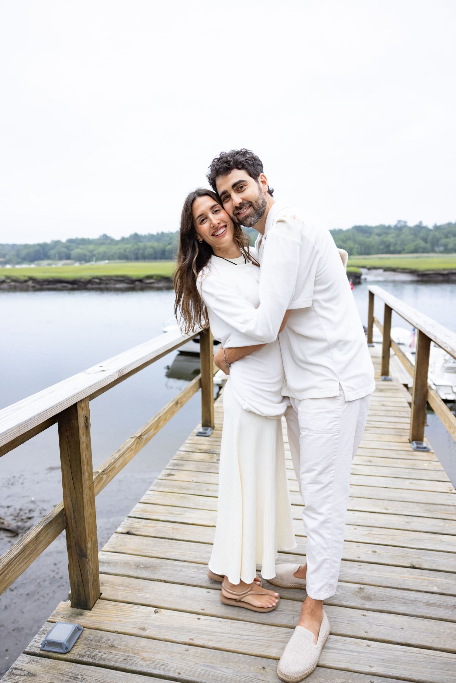 Couple in all white outfits embracing on a wooden dock by the water.