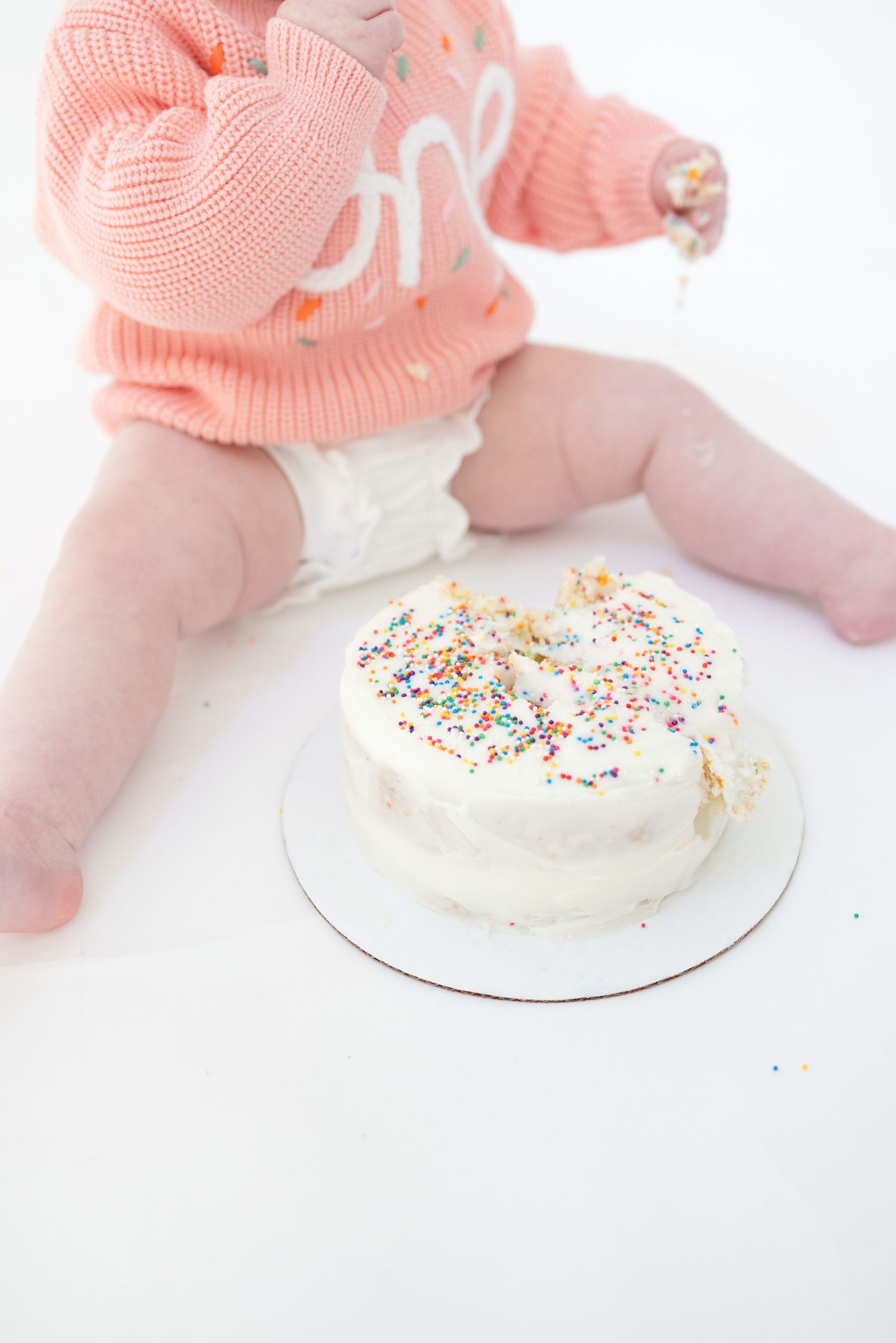 Baby in coral sweater reaching for white cake with sprinkles.