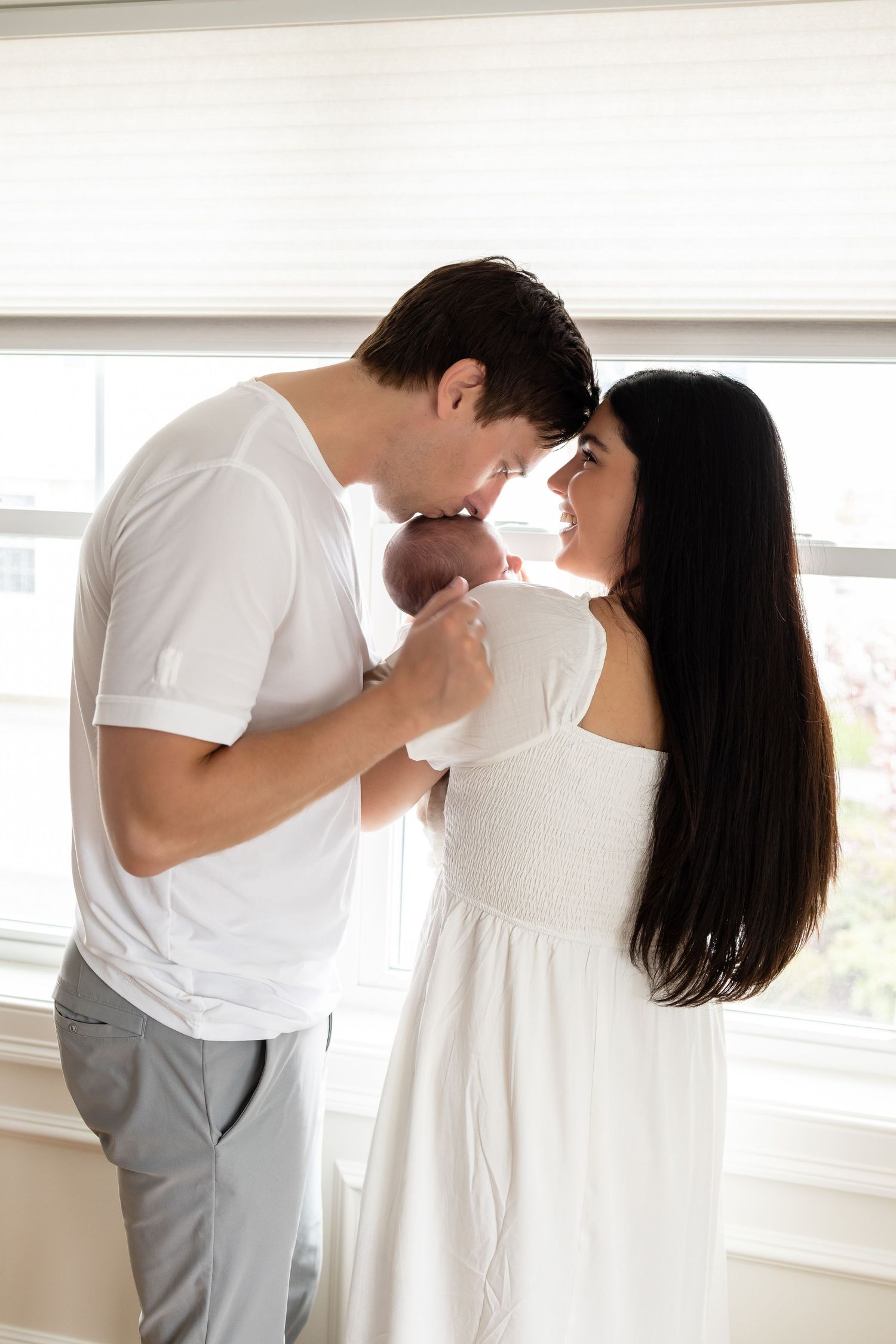A couple in white clothing tenderly kiss their newborn baby near a bright window.