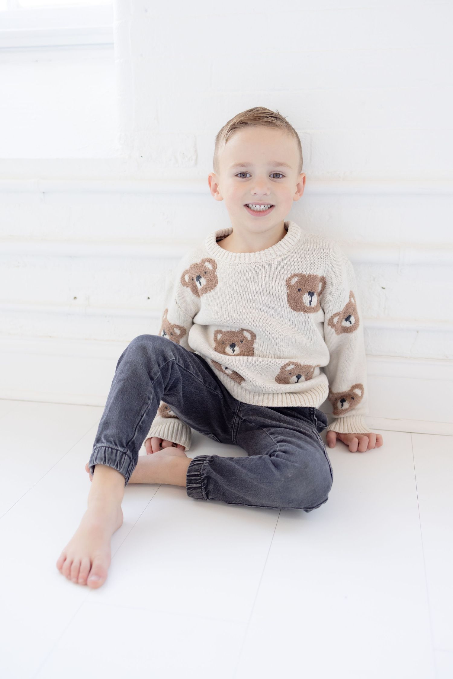 A child sits cross-legged wearing a cream sweater with bear prints in a bright white room.