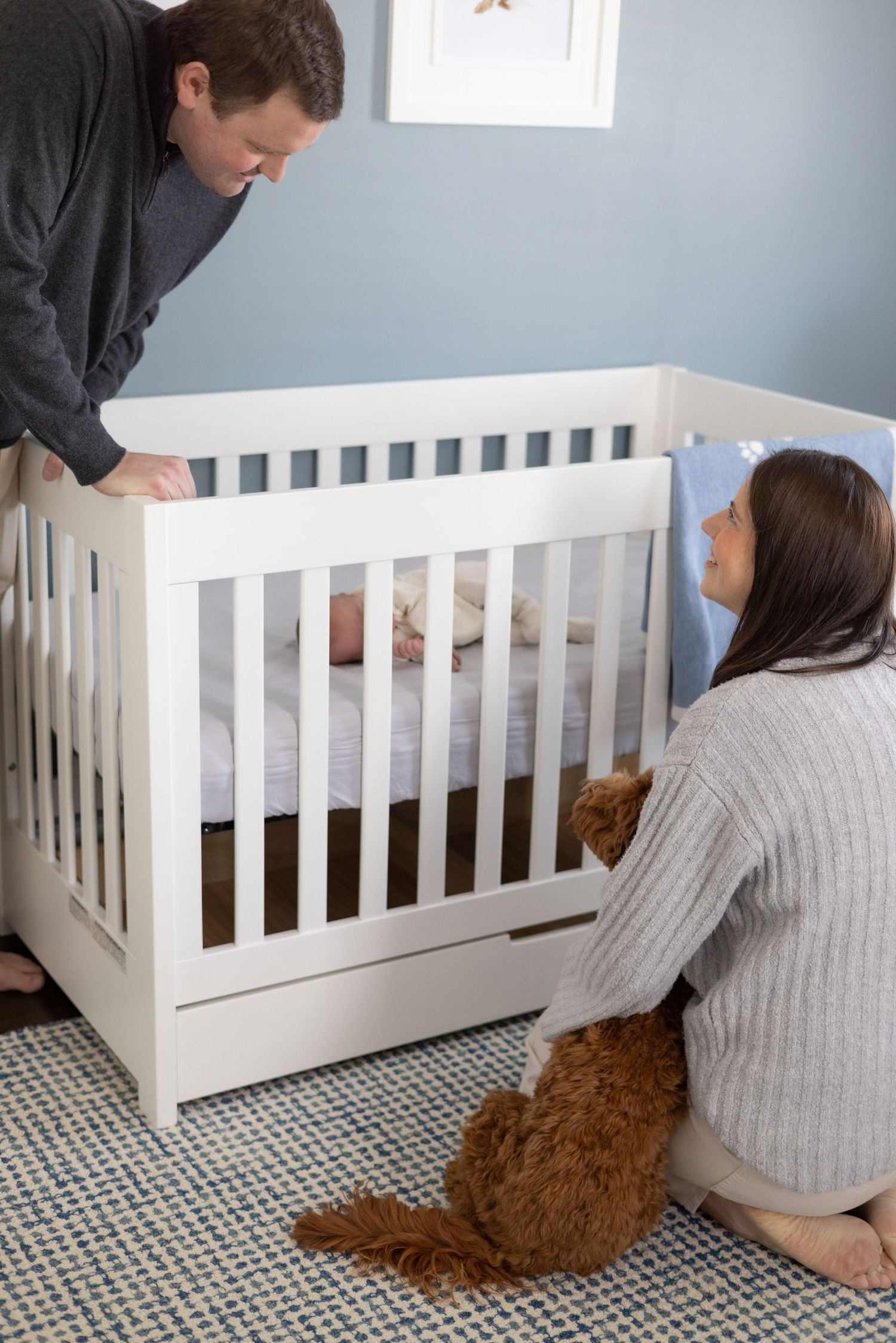 Family and dog gather around white crib in nursery with blue walls.