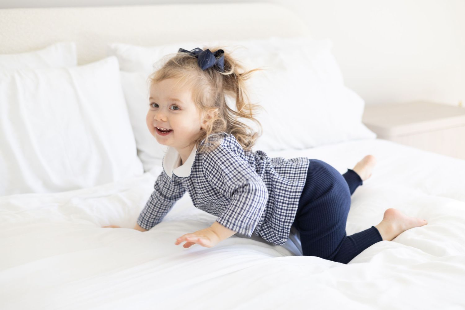 Child in gingham dress playing joyfully on white bedding.