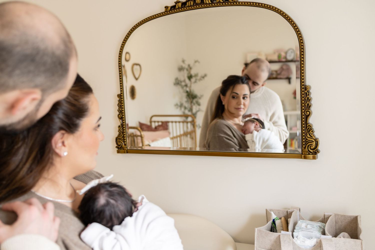 Family with newborn baby reflected in ornate gold mirror in nursery room.