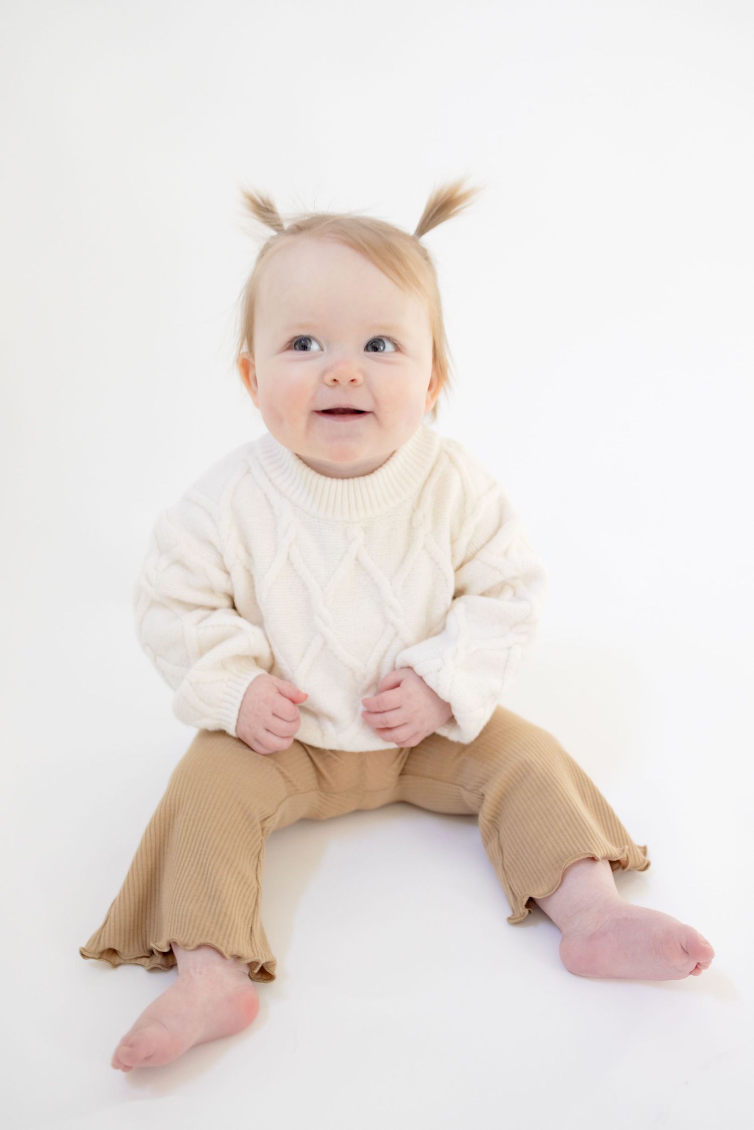 Baby in cream colored outfit sitting against white background.