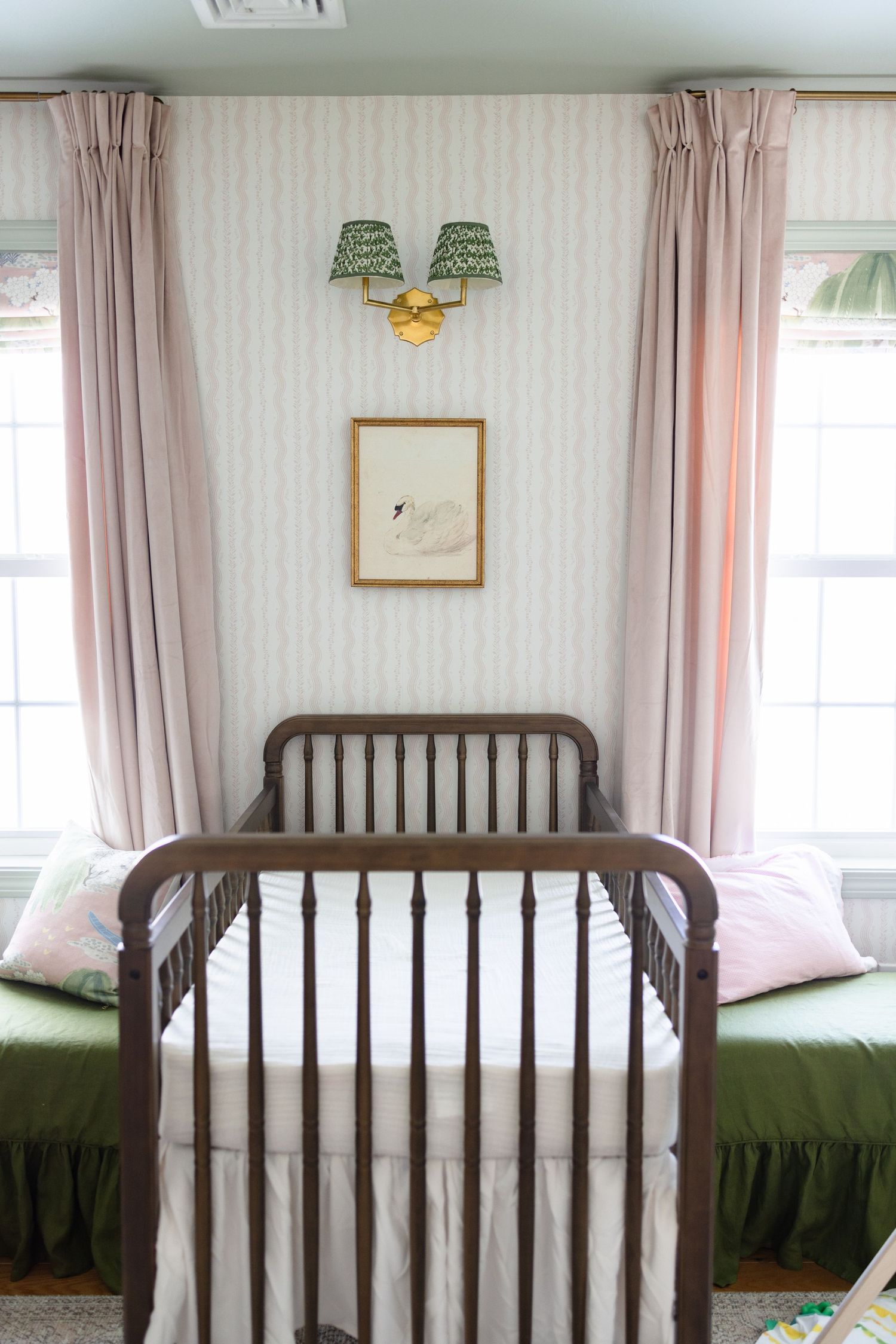 A wooden crib with pink curtains and green bedding sits in a nursery with soft lighting and wall decor.