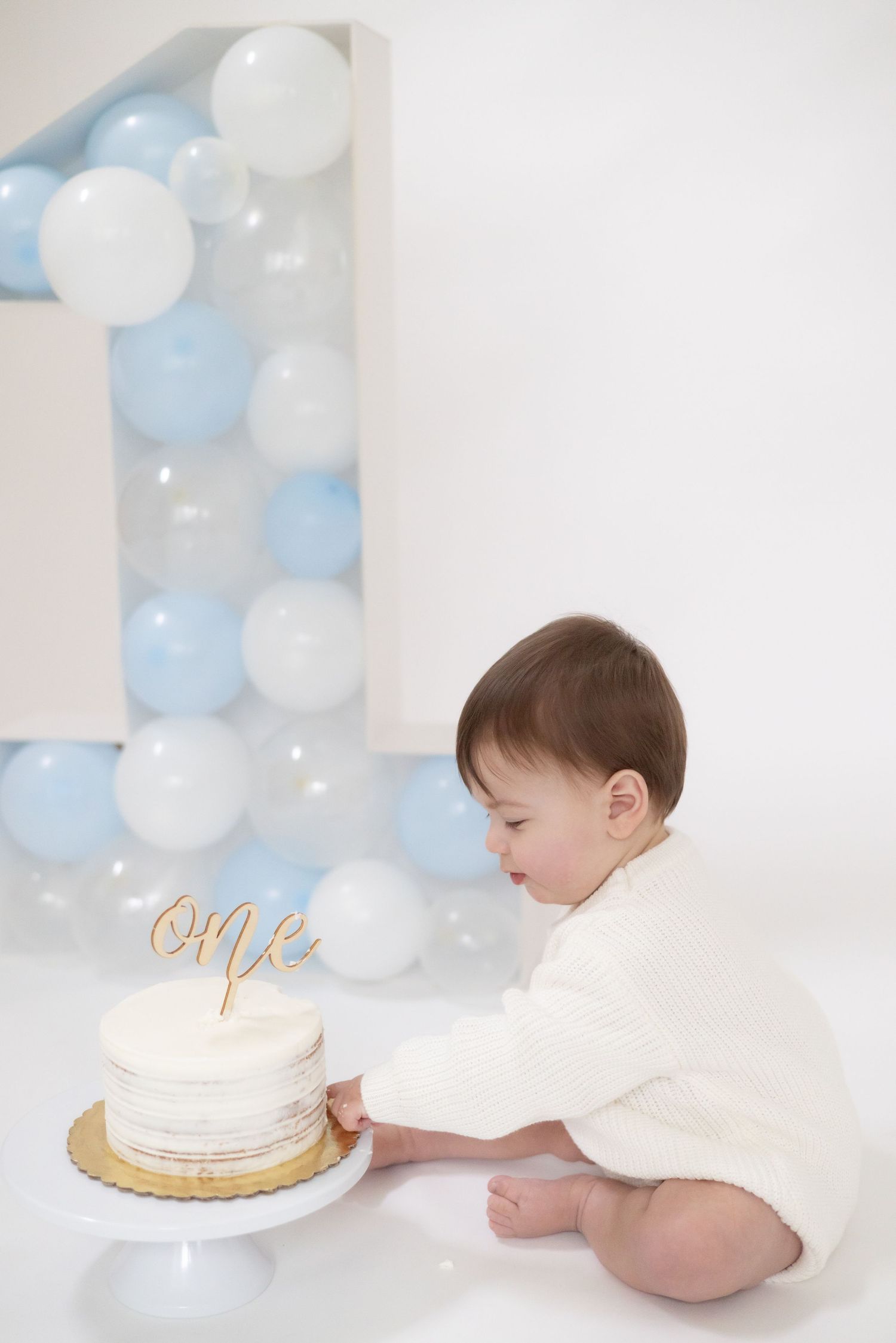Baby in white outfit reaching for birthday cake with blue and white balloon backdrop.