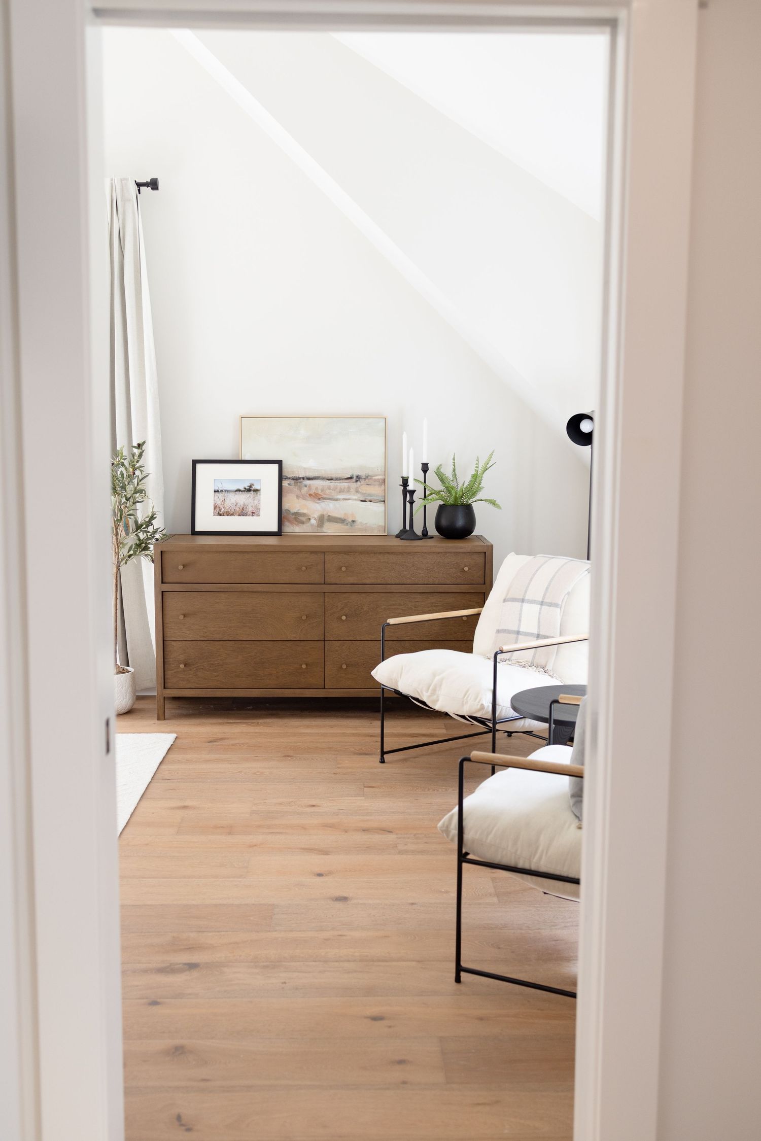 Modern minimalist room with wooden dresser, white chair, and natural light streaming through doorway.