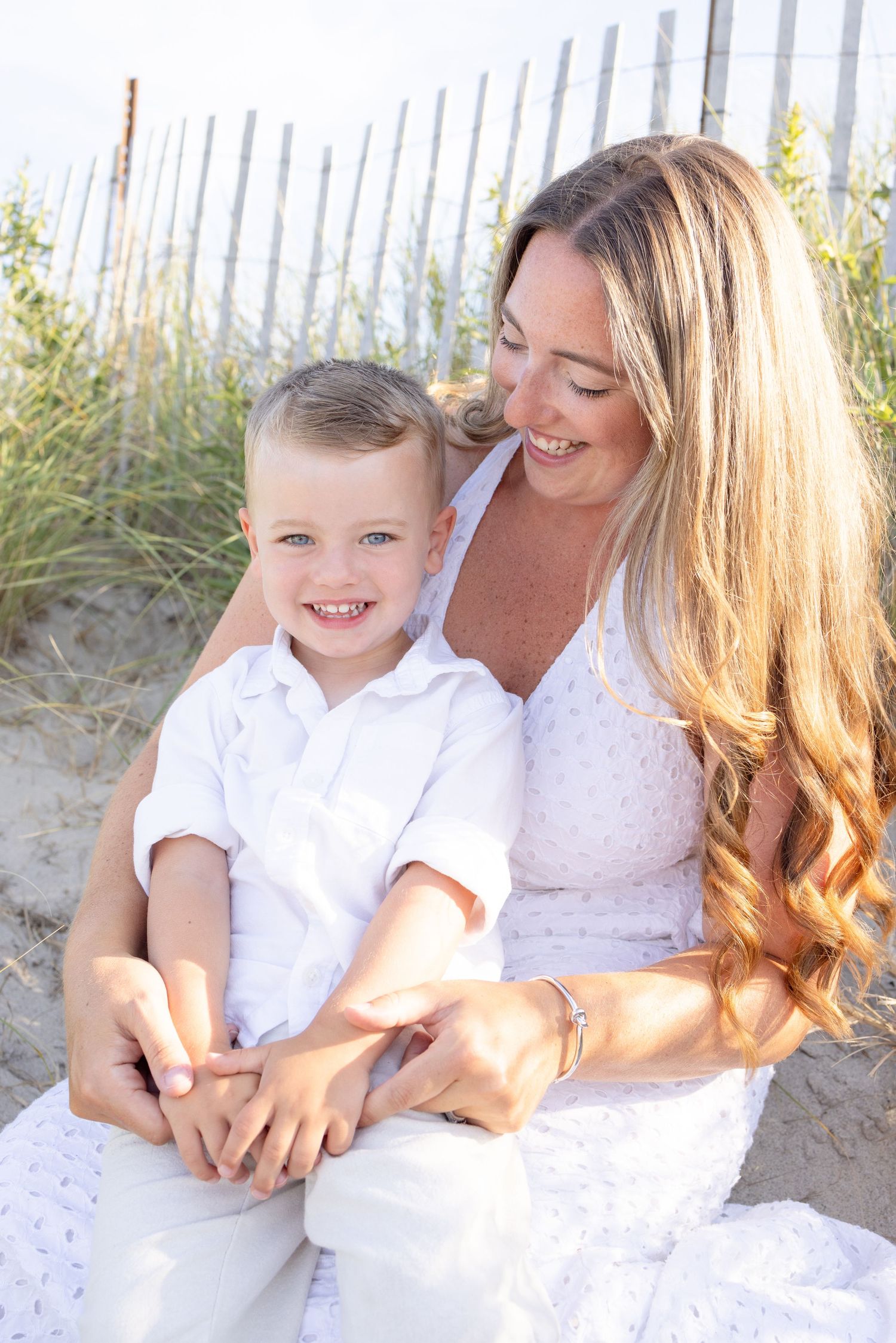 Mother and child sharing tender moment on beach with sand fence in background.