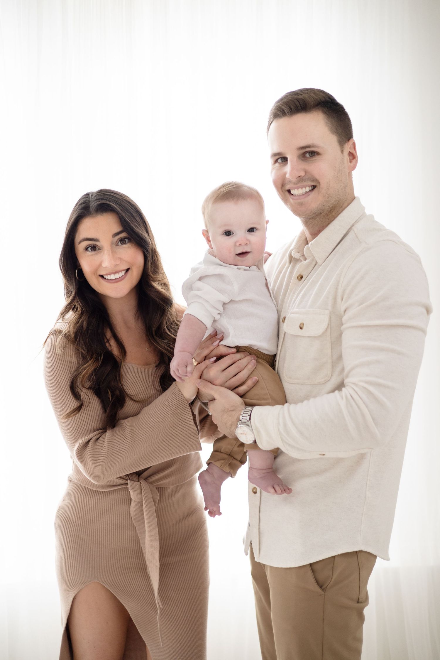 A studio family portrait against white background in coordinating neutral tones.