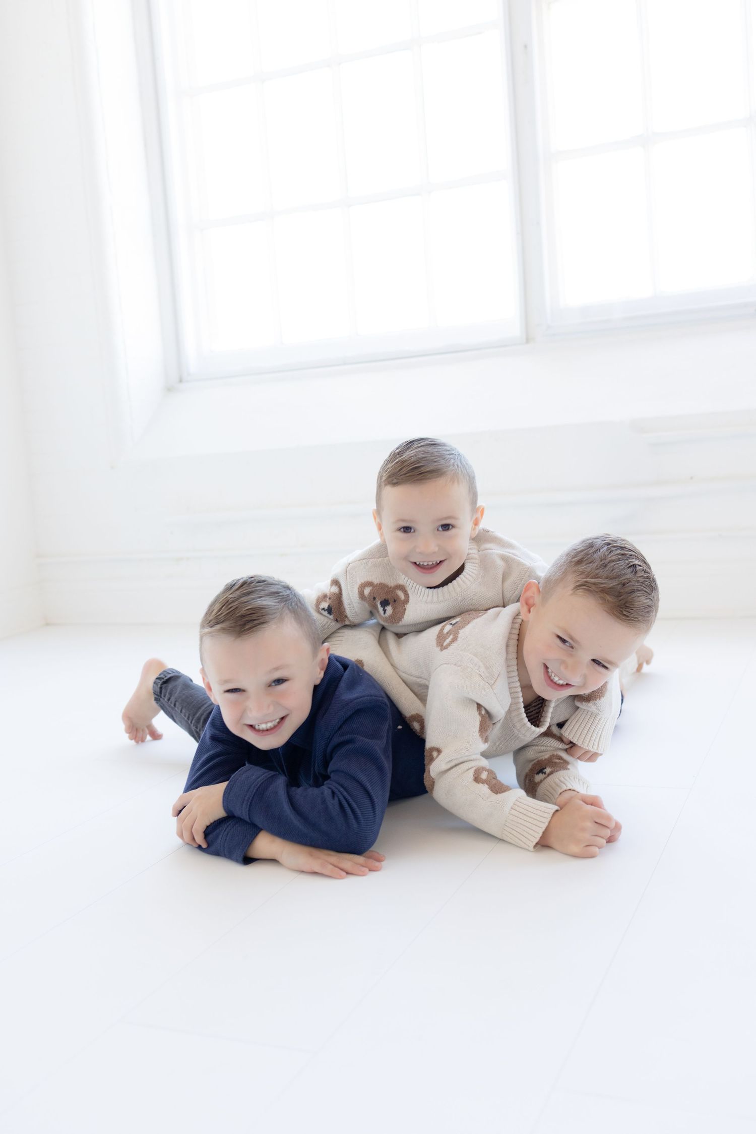 Three children in cozy sweaters playfully pile on top of each other on white floor.