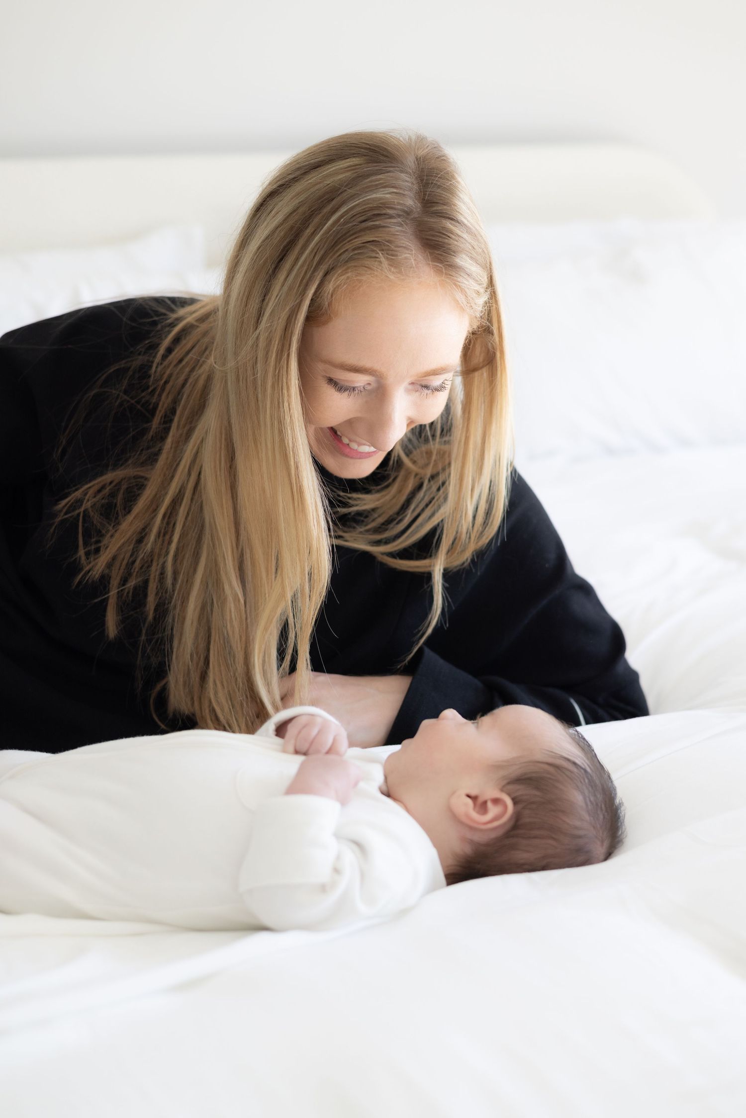 Woman in black sweater lovingly gazes at baby on white bedding.