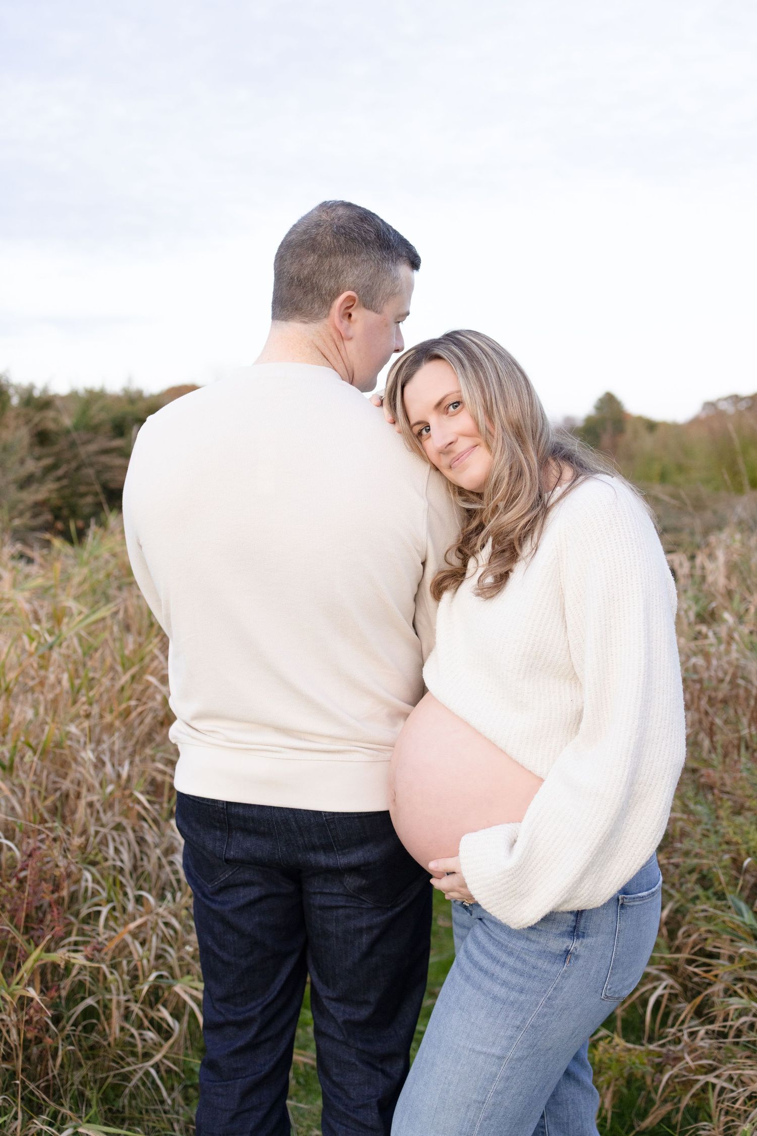 Expectant couple in cream sweaters embracing outdoors among tall grasses.