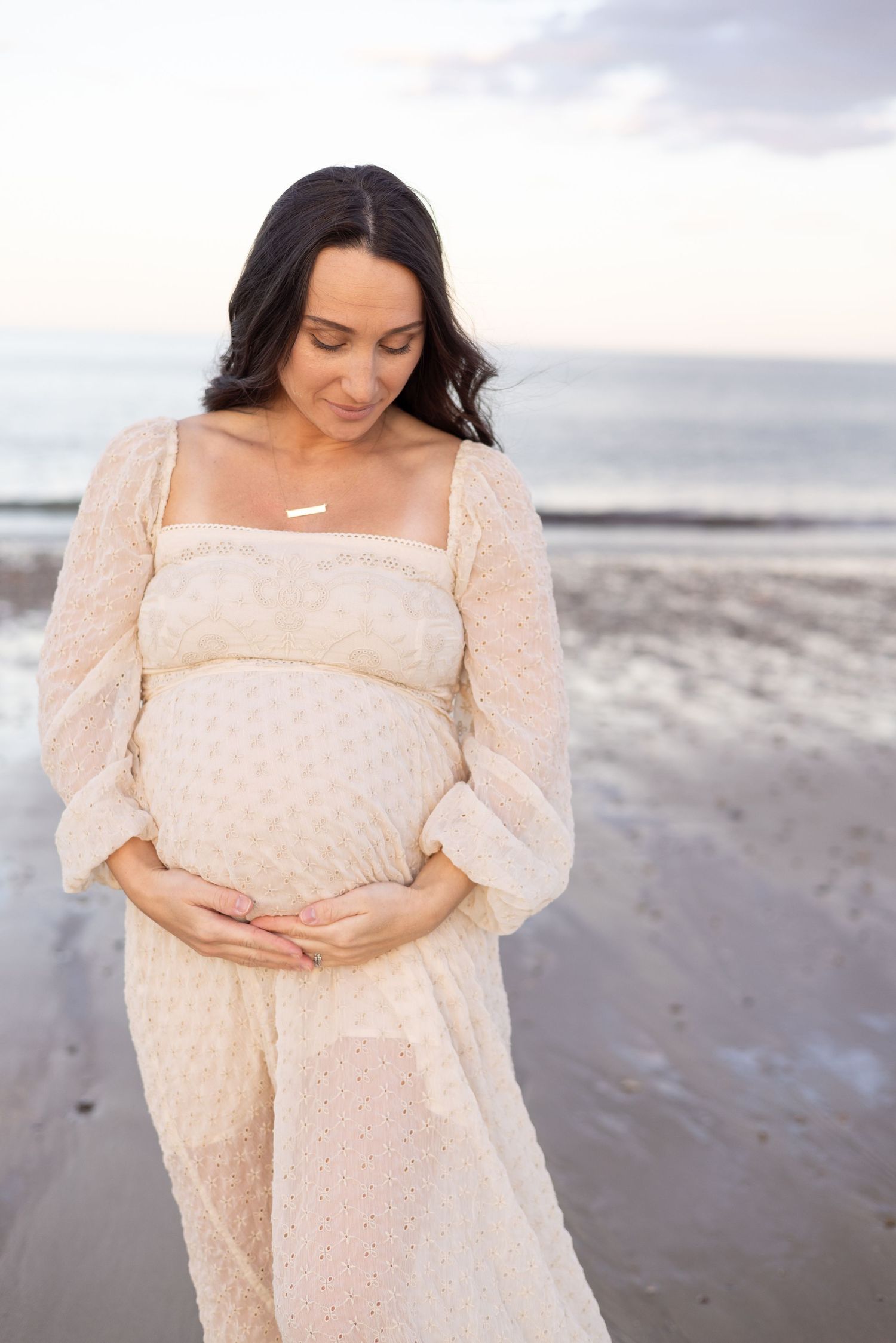 Maternity photos on beach featuring woman in cream dotted dress at sunset.