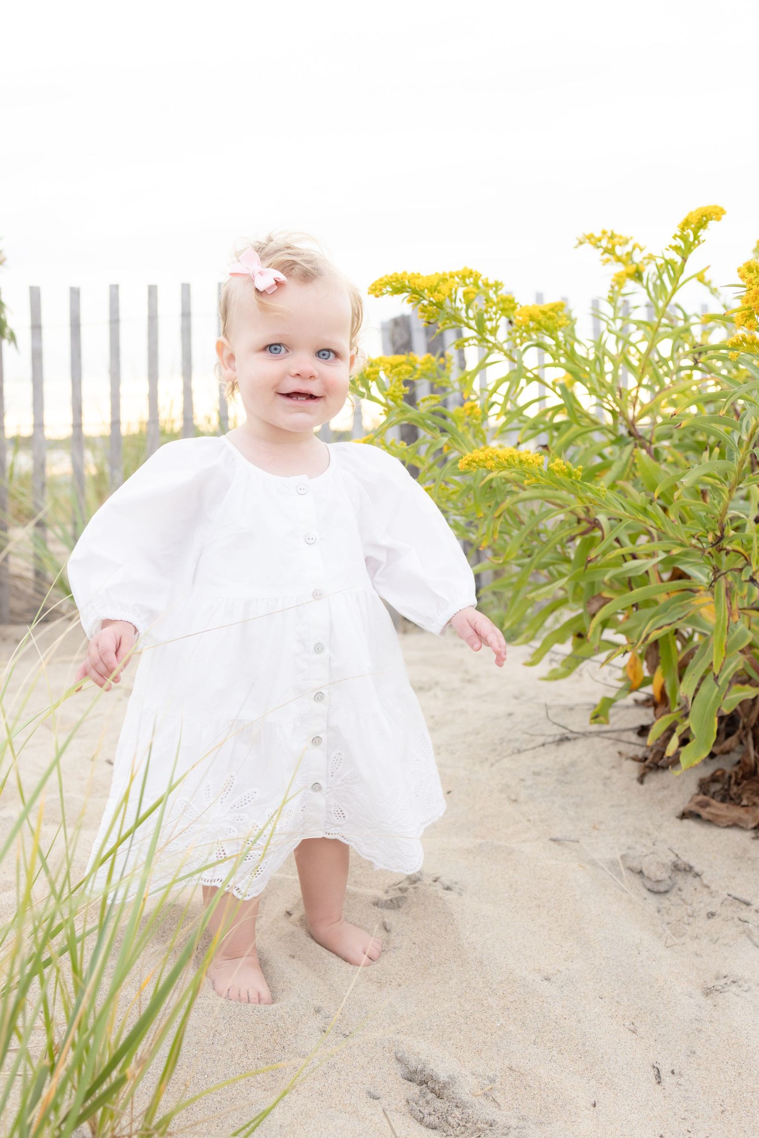 Baby in white dress playing in beach sand near coastal plants.
