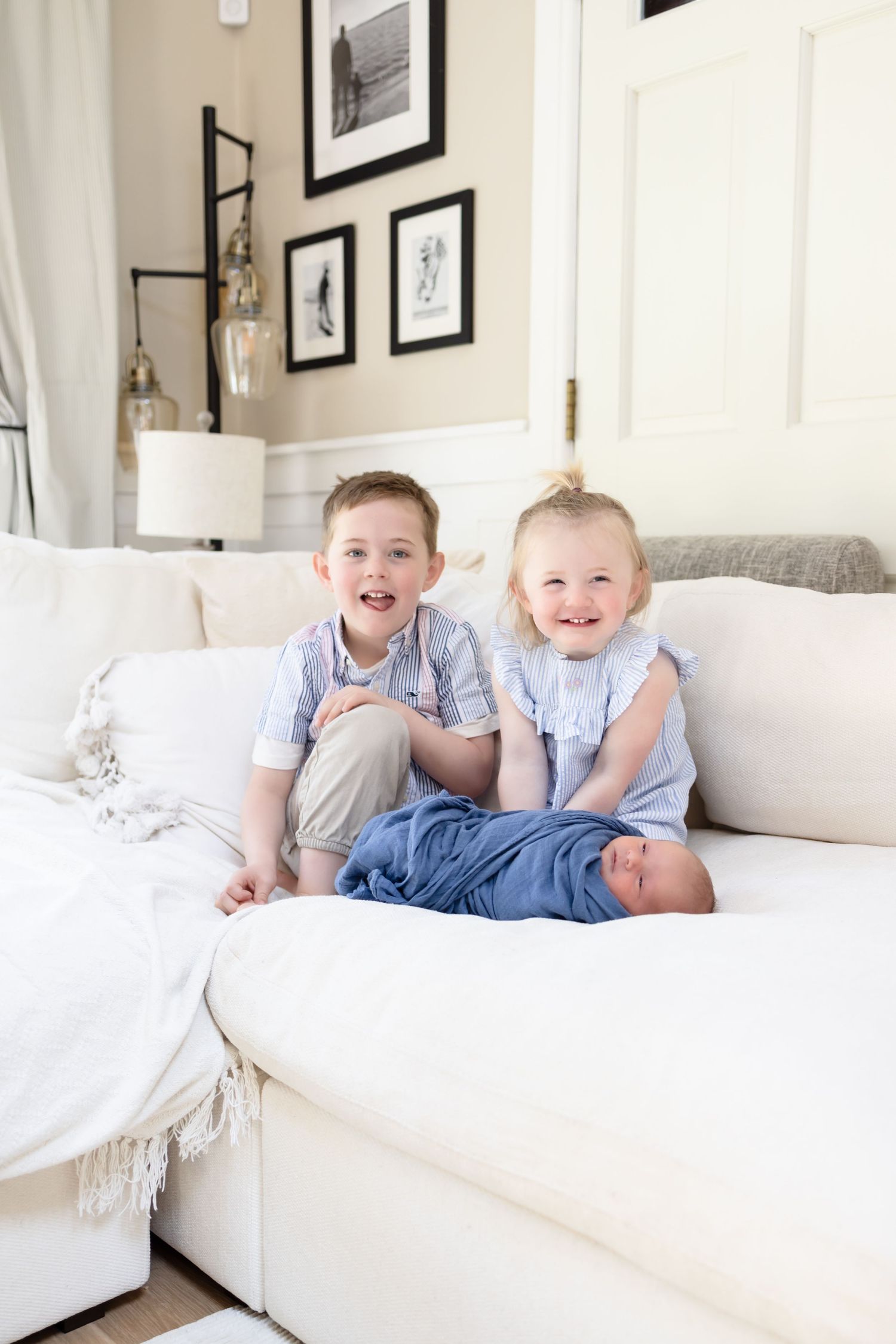 Two smiling children sit on a white sofa with a swaddled newborn baby between them in a bright living room.