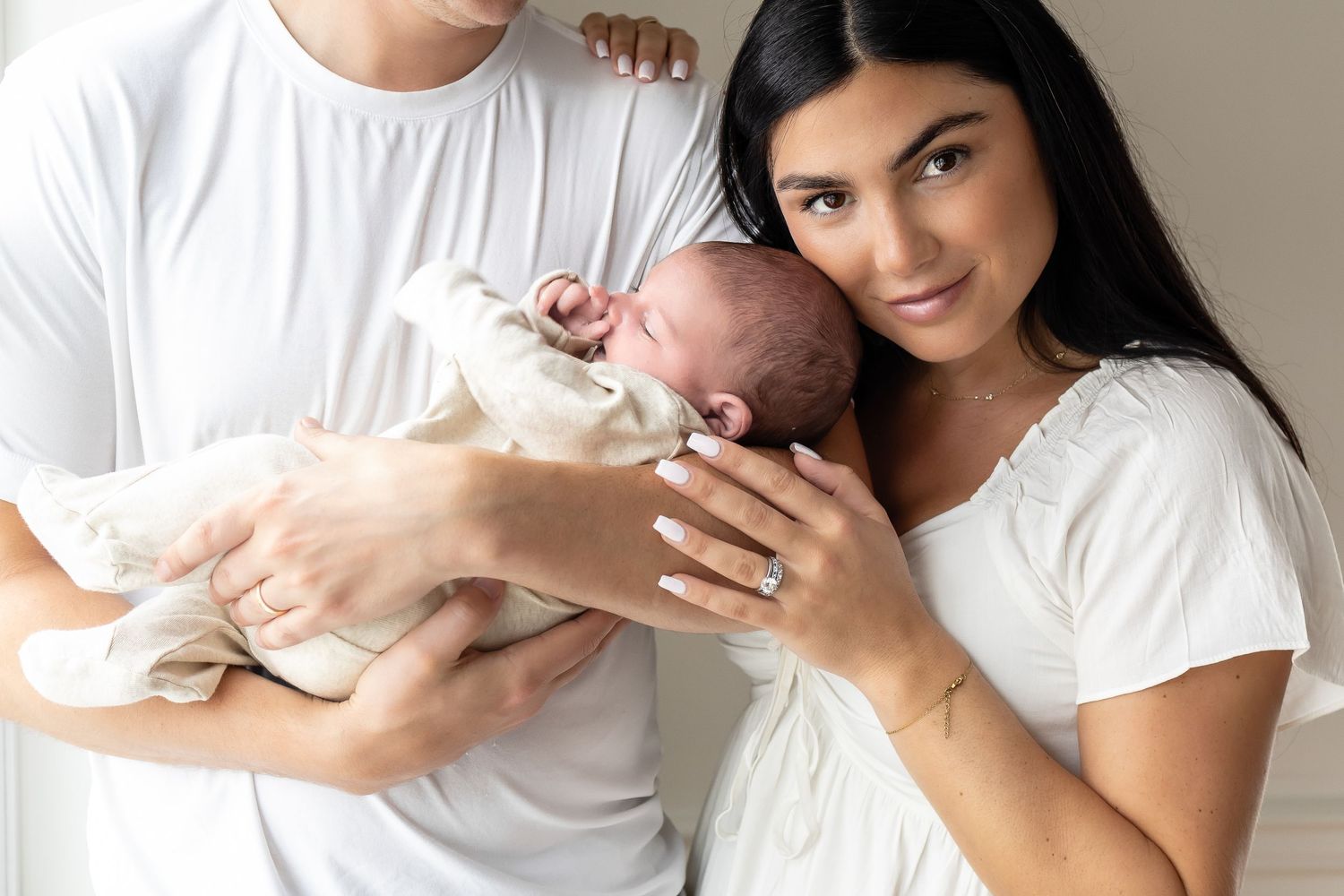 A couple in white outfits tenderly holds their newborn baby, smiling warmly in a bright, softly lit room.