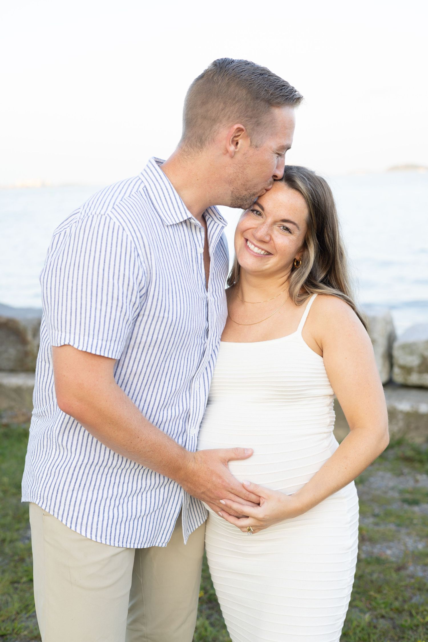Couple embraces on a beach in coordinated white and striped outfits during a romantic photoshoot.