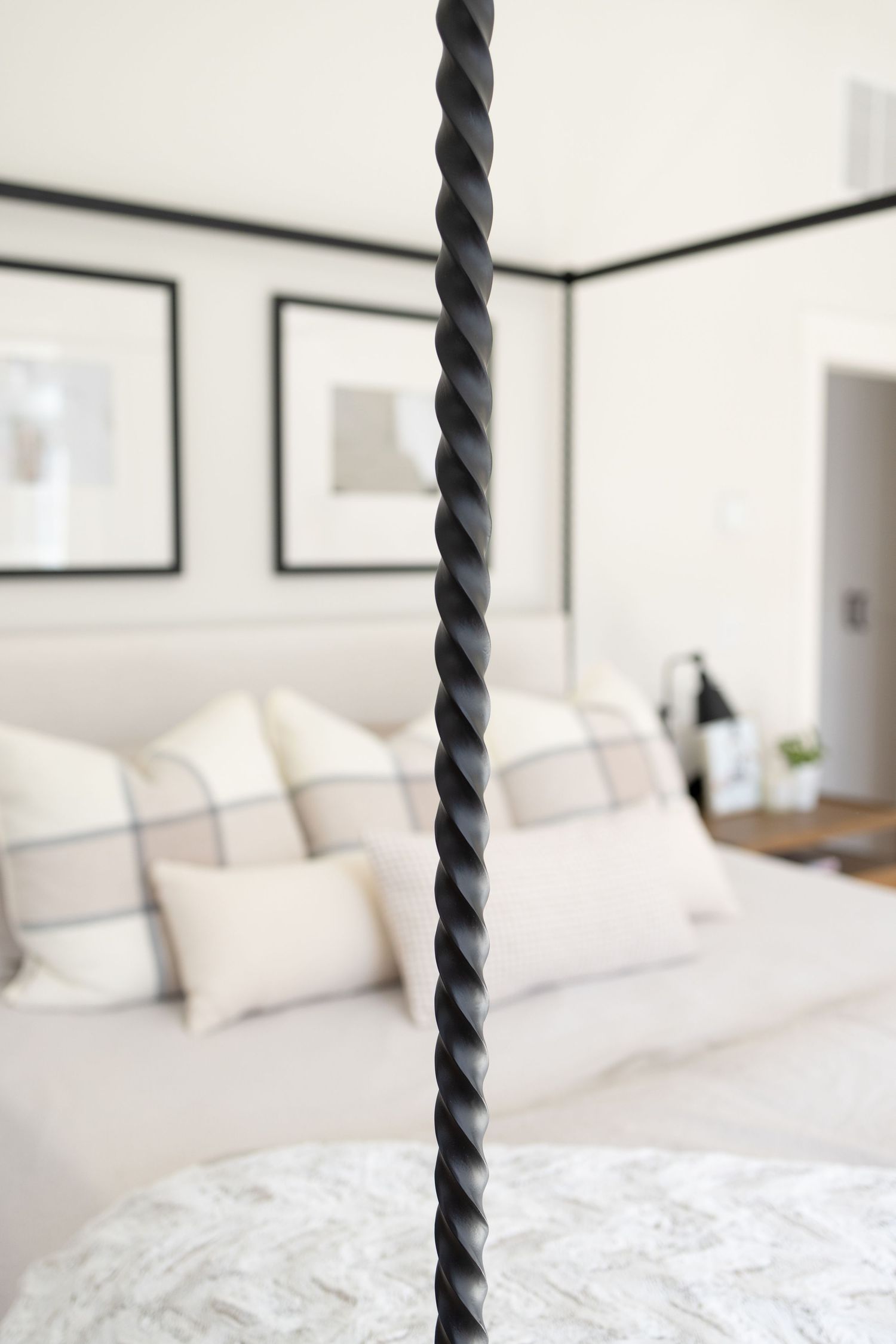 Close-up of a black twisted rope post against a white bedroom with plush pillows and framed artwork.