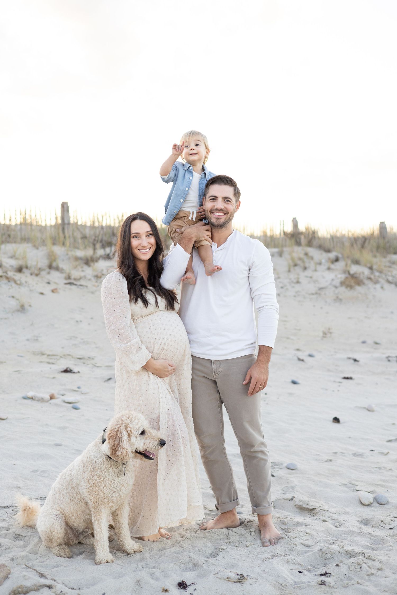 Family portrait on beach with white dog and sand dunes in background.