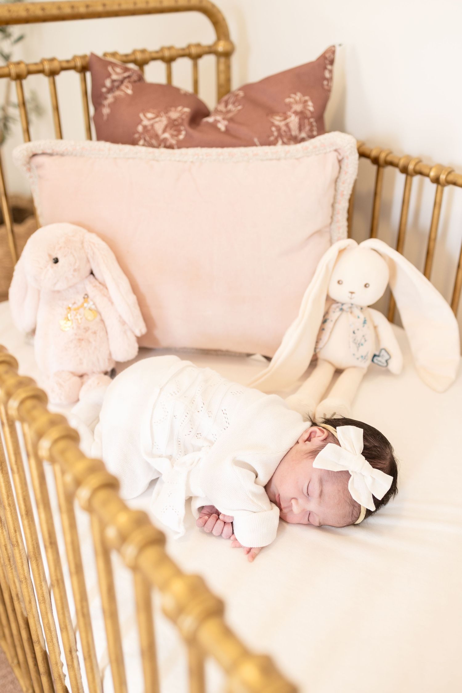 Newborn baby sleeping in gold crib with white bow headband, surrounded by bunny stuffed animals and pink pillows.