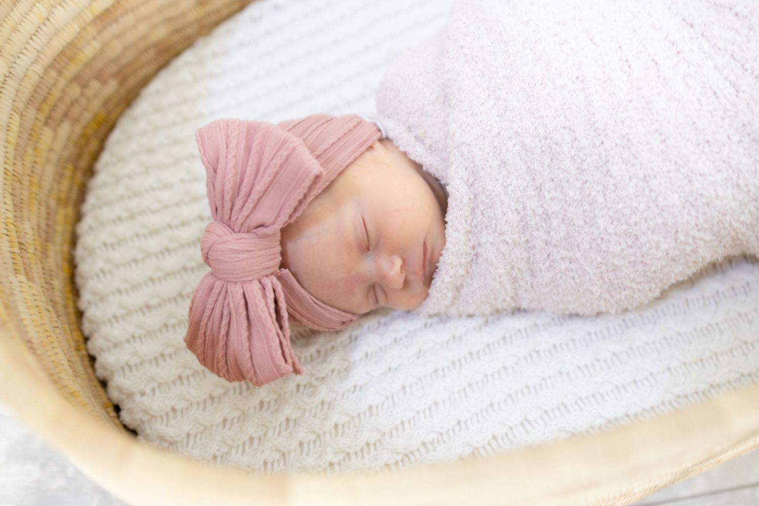 Newborn baby sleeping peacefully in white bassinet with pink swaddle.