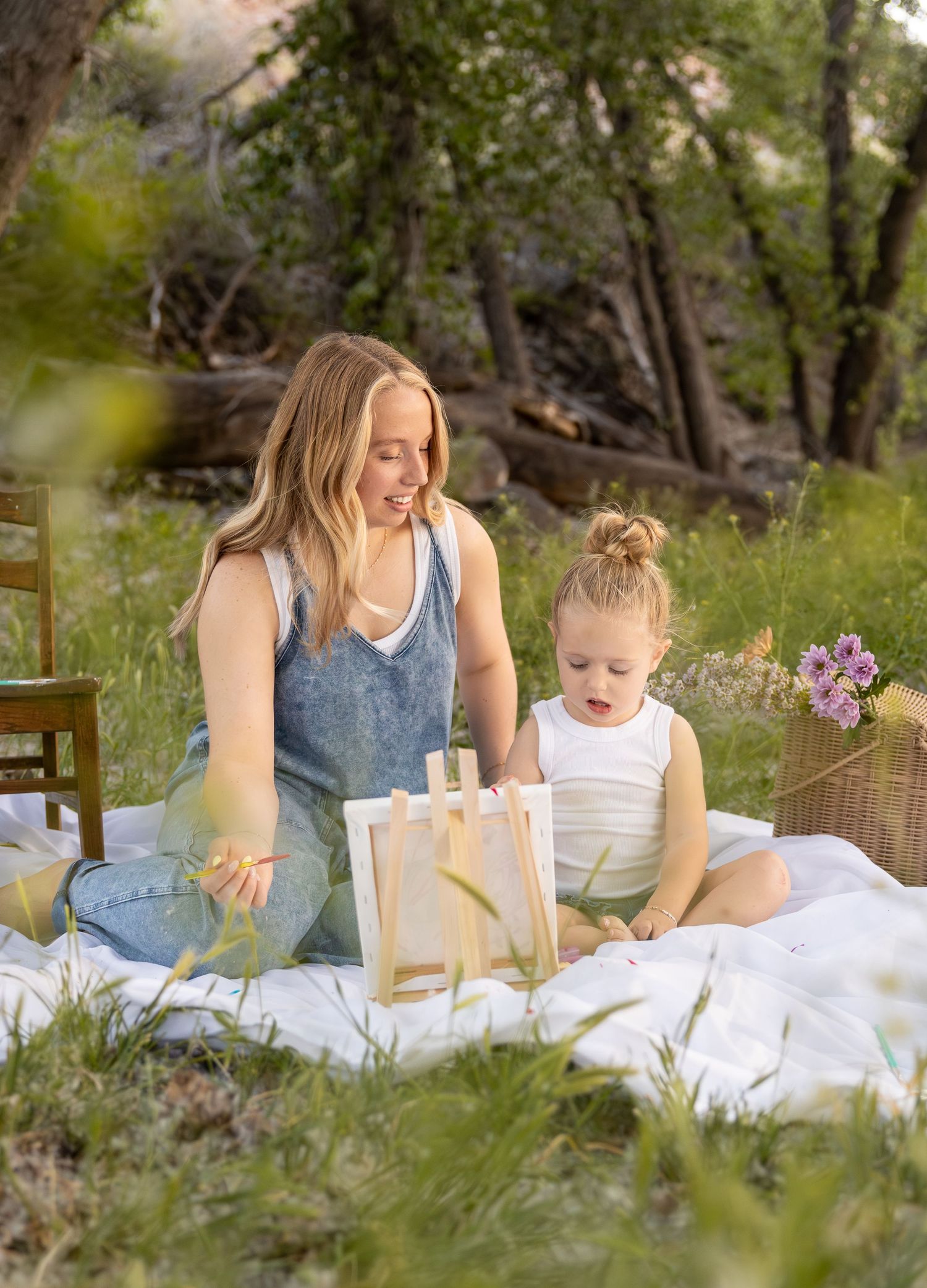 A mom and her daughter are sitting in a field painting a canvas together.