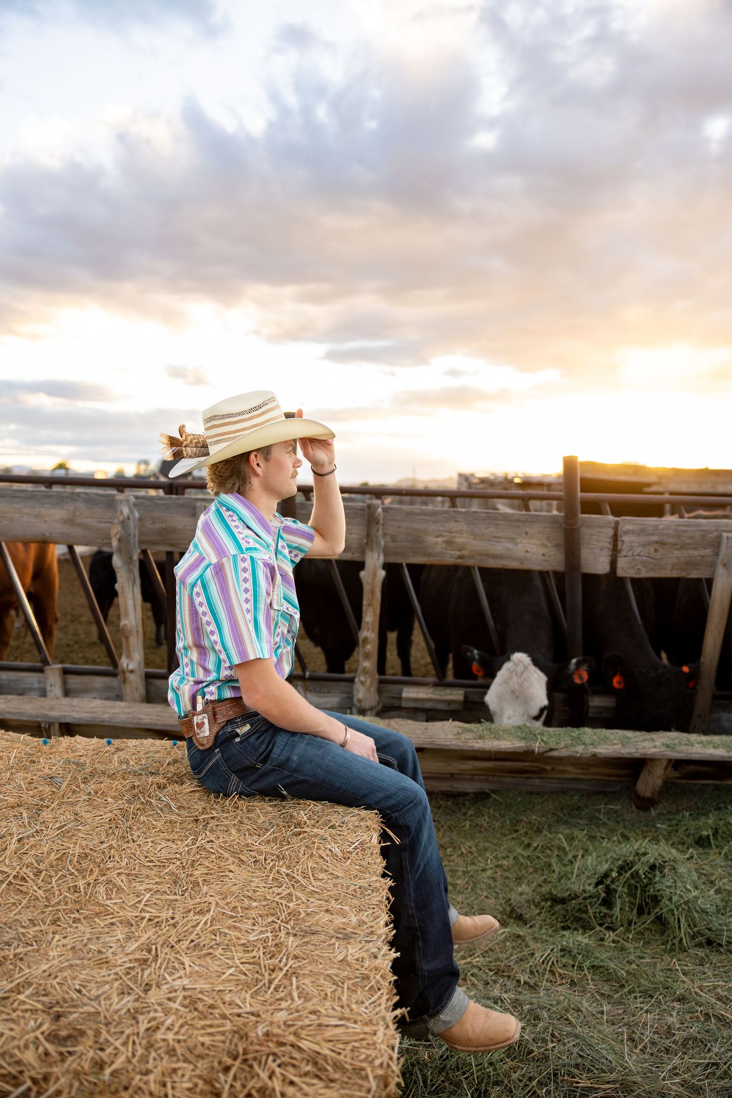A senior boy is sitting on a hay bale holding his cowboy hat as he stares into the sunset on his family farm.