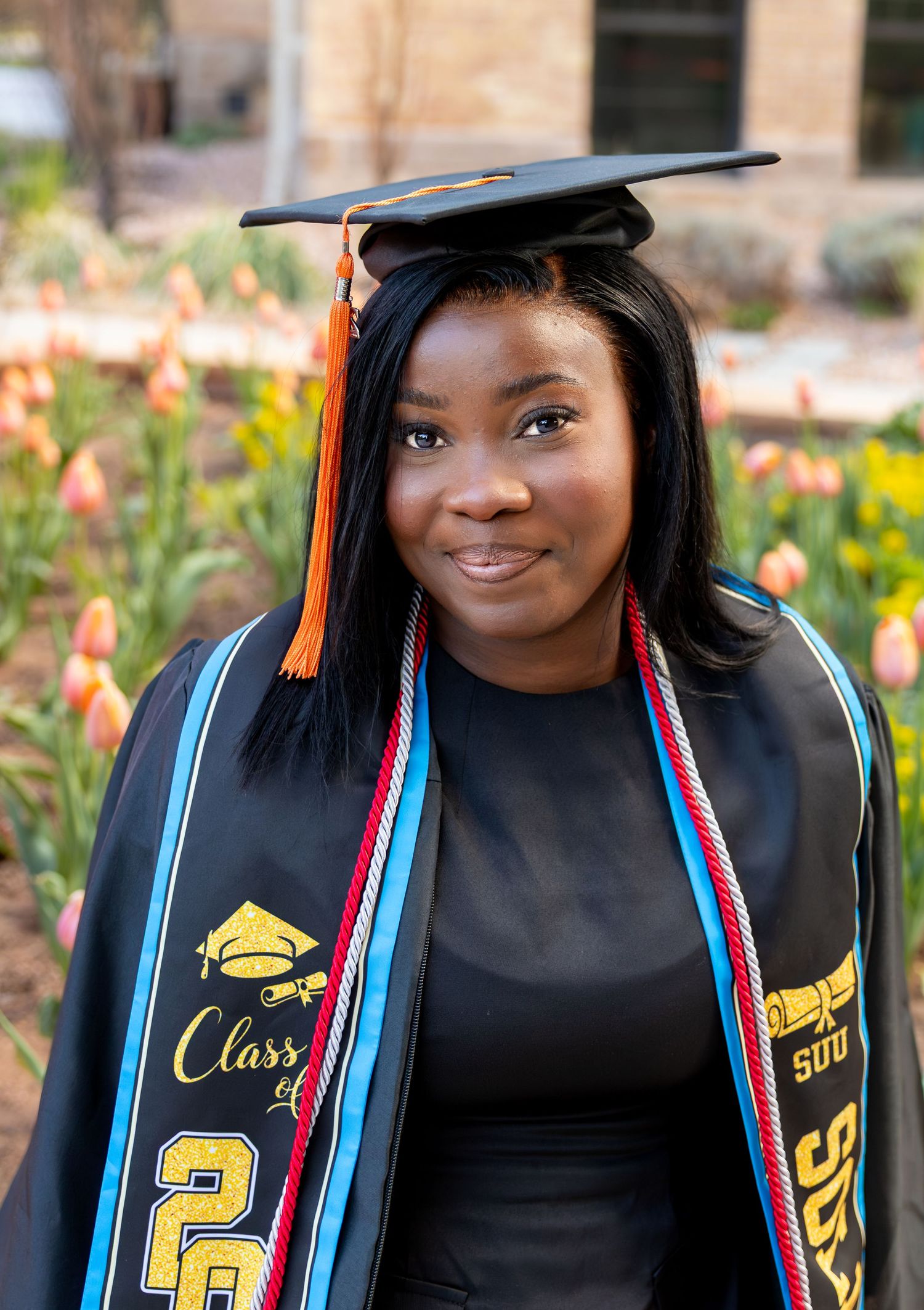 A college graduate is sitting in front of a field of flowers with her cap and gown on.