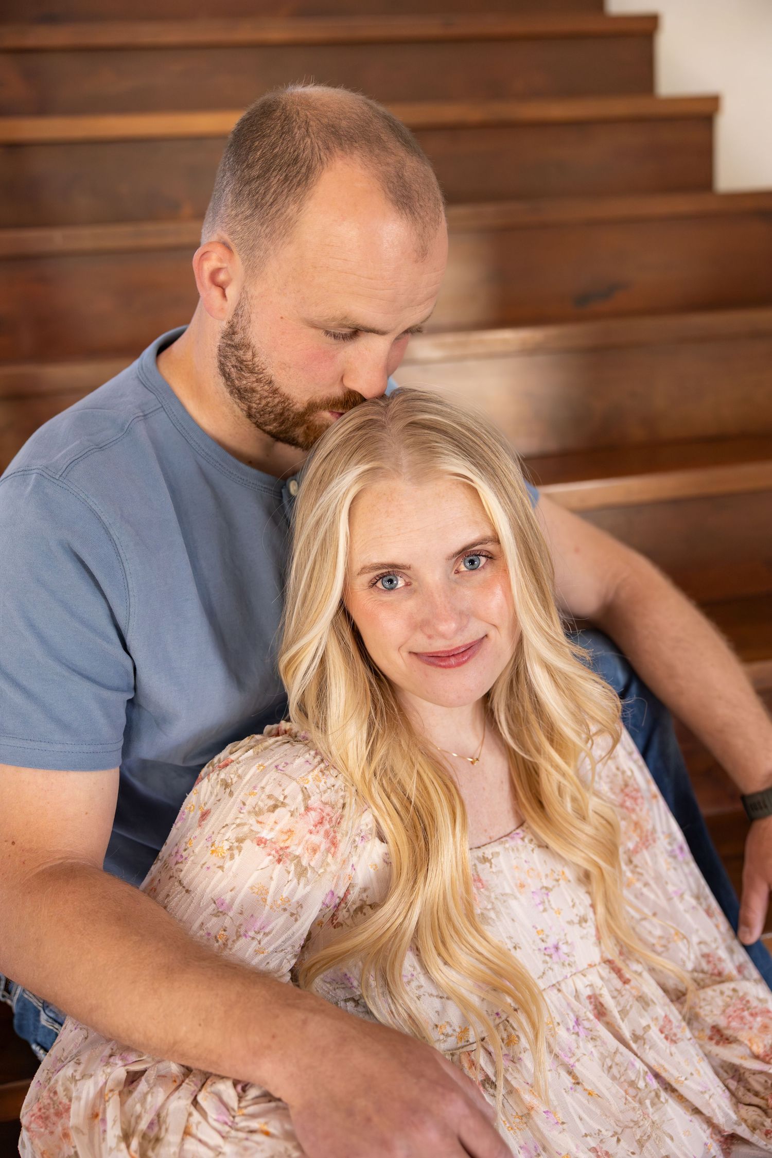A couple are sitting on some stairs. The husband is kissing his wife on the top of her head.